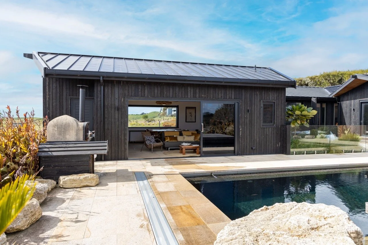 Modern black wooden house with a metal roof, large sliding glass door, outdoor seating, and a swimming pool with rocks and plants in a scenic landscape.