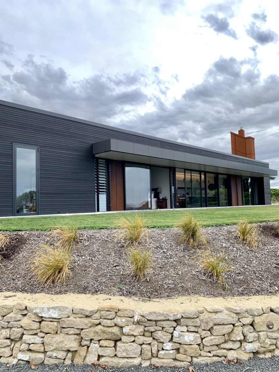 Modern house with large glass windows, dark exterior siding, a brown brick chimney, and a landscaped yard with grass and small bushes.