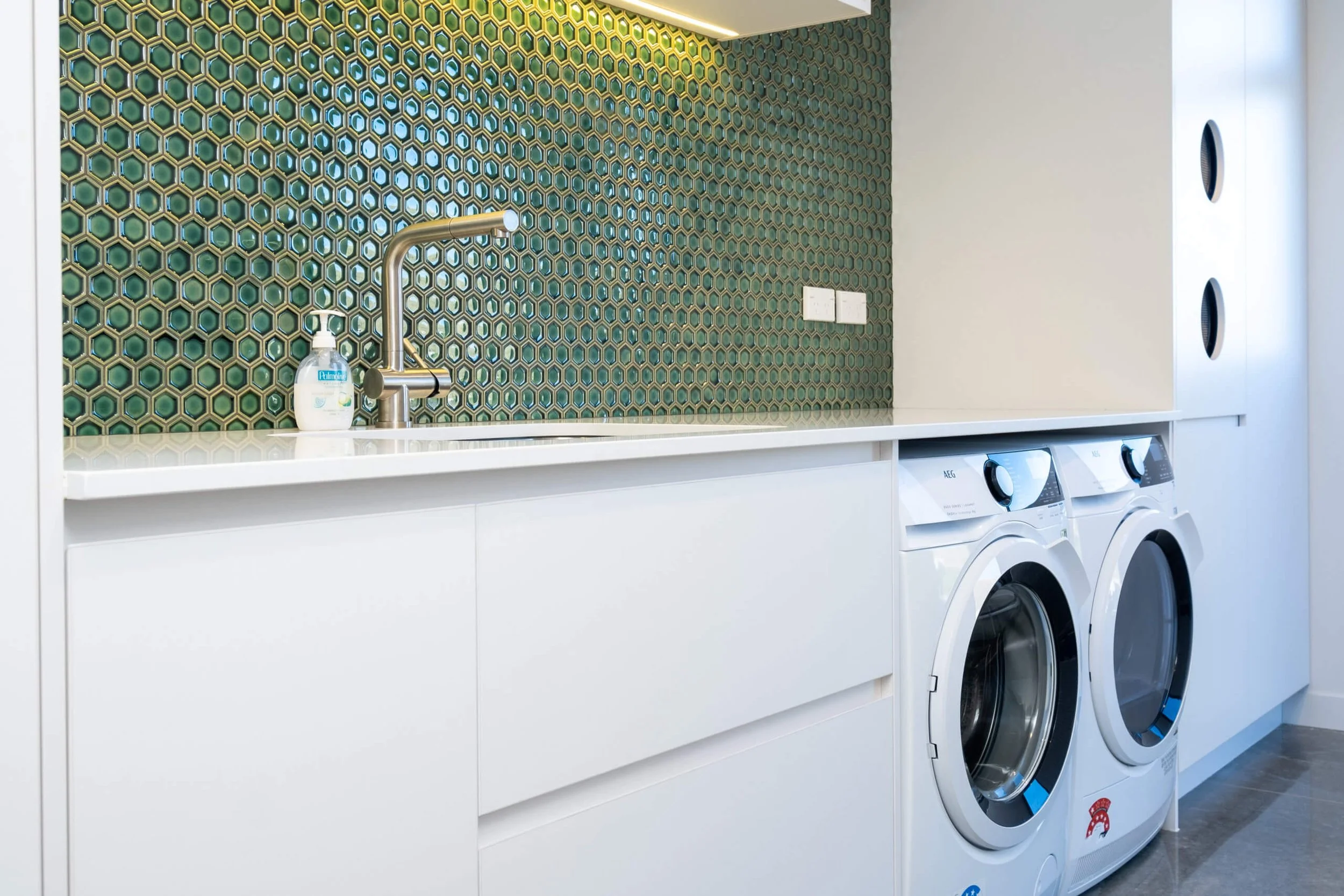 A modern laundry room with white cabinetry, a sink, and a patterned green and brown tile backsplash. There are two front-loading washing machines and a soap dispenser on the counter.