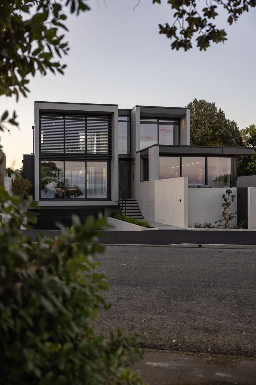 Modern multi-story house with large glass windows and a minimalist design, surrounded by trees and a paved road