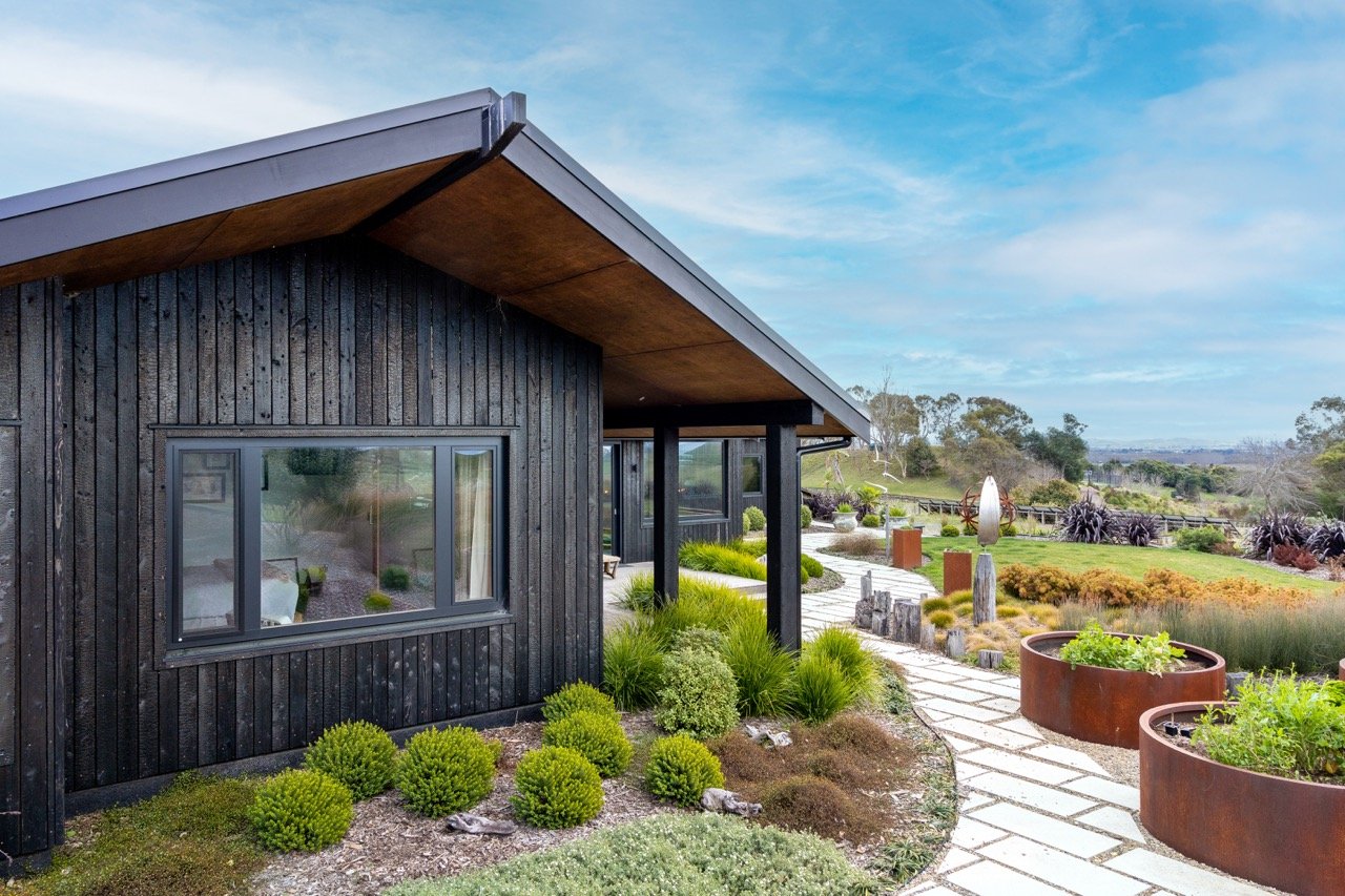Modern house with dark wooden exterior and a stone pathway leading through landscaped garden with plants and decorative features