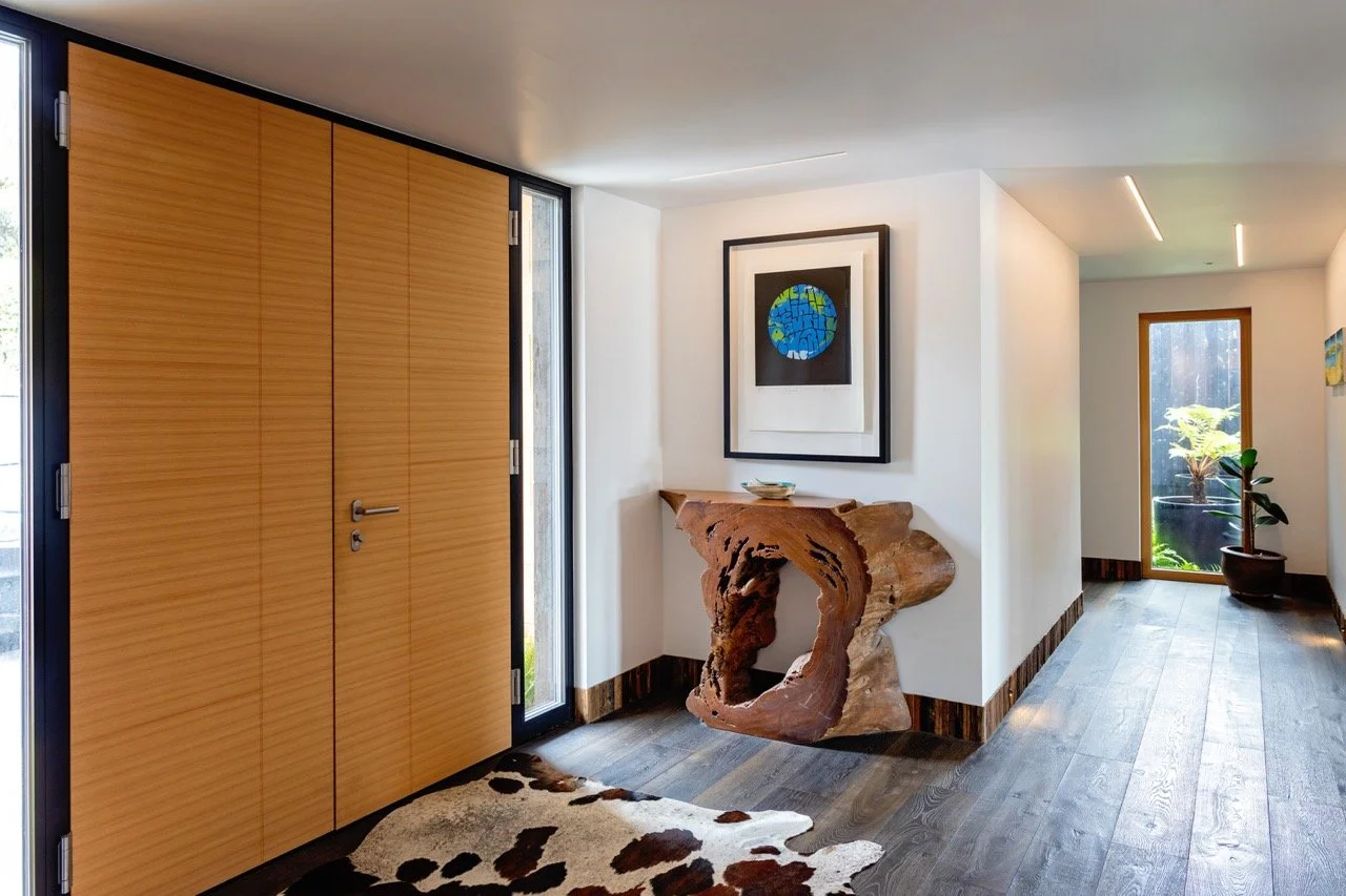 Modern interior entryway with wooden door, abstract art, decorative wood sculpture, potted plant, and cowhide rug.