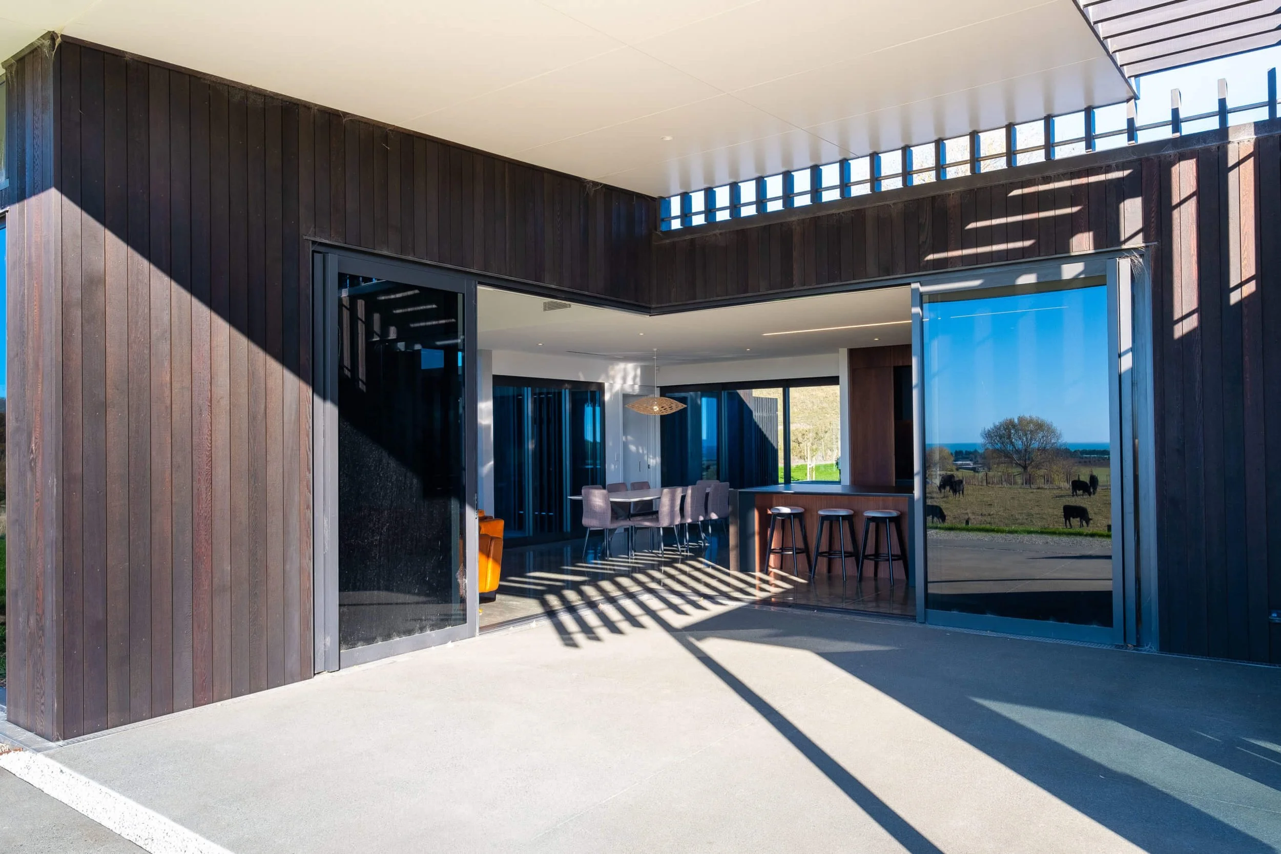 Modern house with large sliding glass doors open to patio, view of grassy field with grazing horses, and blue sky in background.