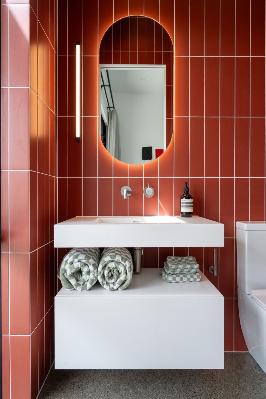 Modern bathroom with red tiled walls, a white rectangular sink with folded towels underneath, an oval backlit mirror, and a liquid soap dispenser on the sink.