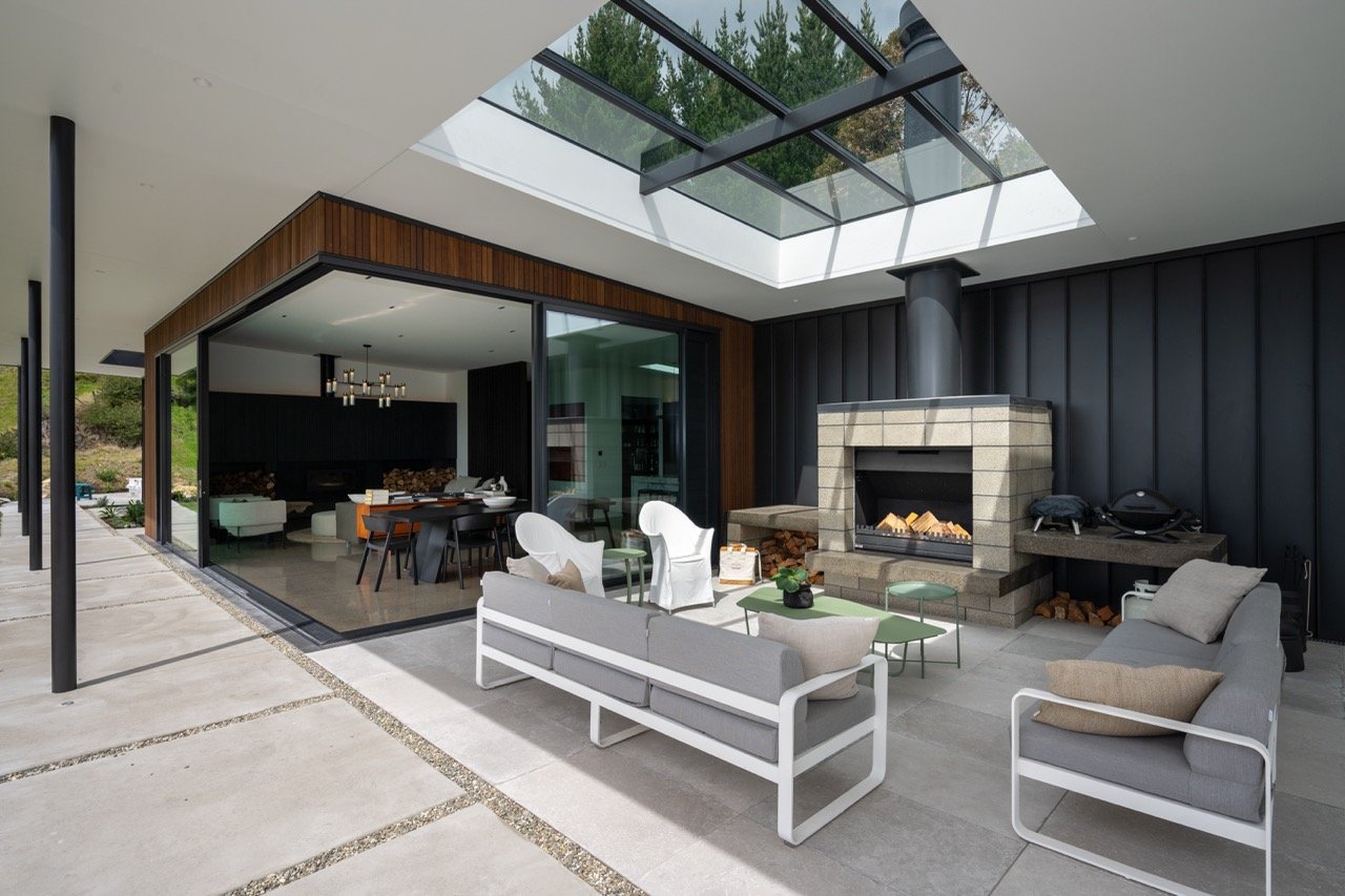 Outdoor patio area with seating, a fireplace with logs, and a large skylight above, adjoining a modern interior dining and living space.