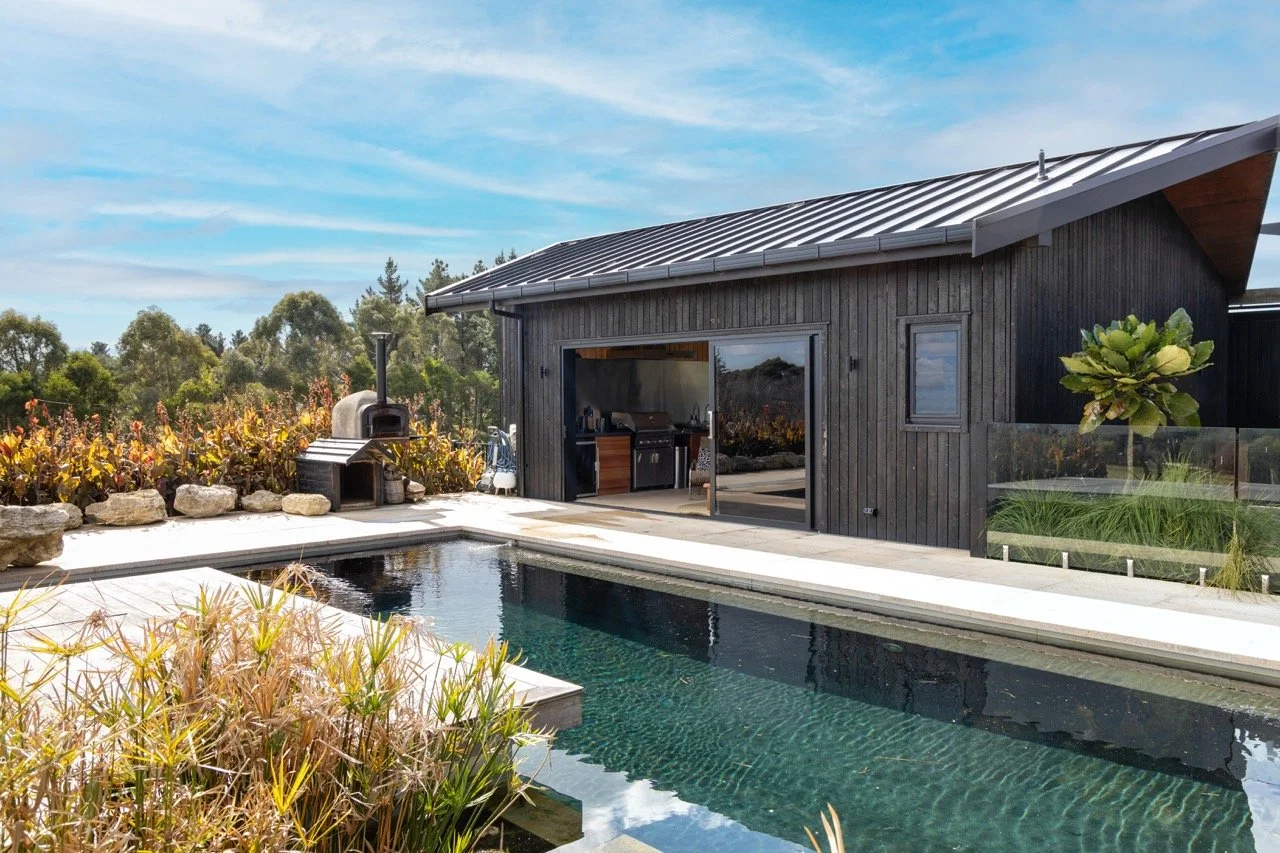 A backyard with a swimming pool in front of a modern black wooden house. The house has an open door leading to an outdoor kitchen area. There is a wood-fired oven on the deck and lush landscaping with various plants and trees around.