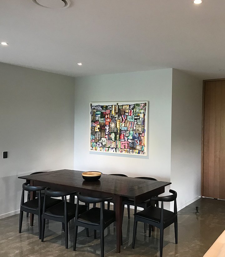 A dining room with a dark wooden table surrounded by six black chairs, a decorative bowl on the table, a colorful abstract painting on a white wall, and a wooden door on the right side.