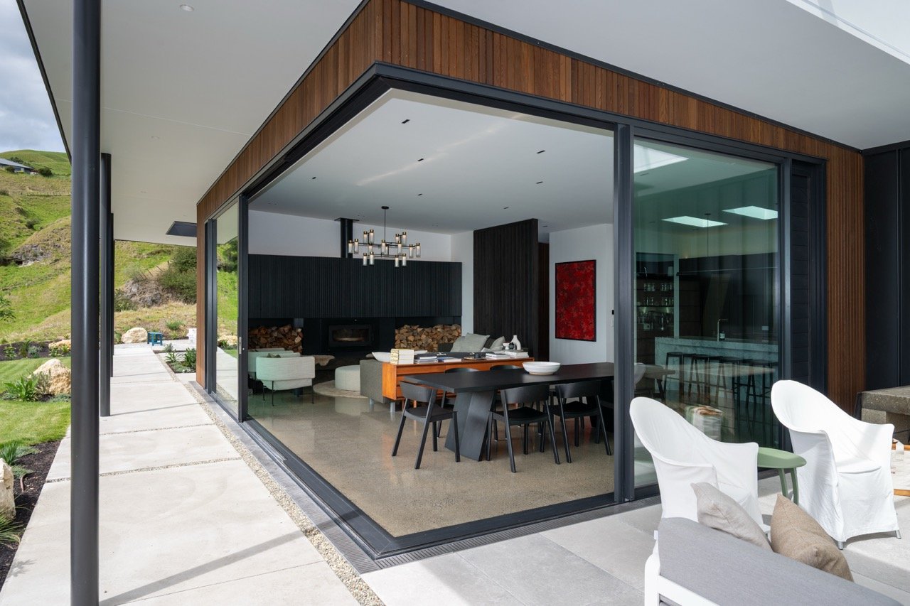 Modern home with large glass sliding doors opening to a patio, showing an interior living and dining area with contemporary furniture and decor, surrounded by a landscaped yard and hills.