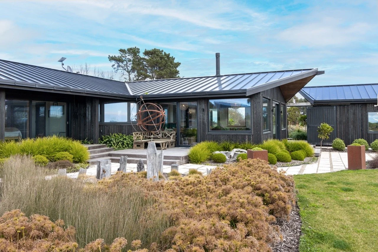 Modern black house with metal roof and large windows, surrounded by landscaped garden with various shrubs, grasses, and stone pathway, under a partly cloudy sky.