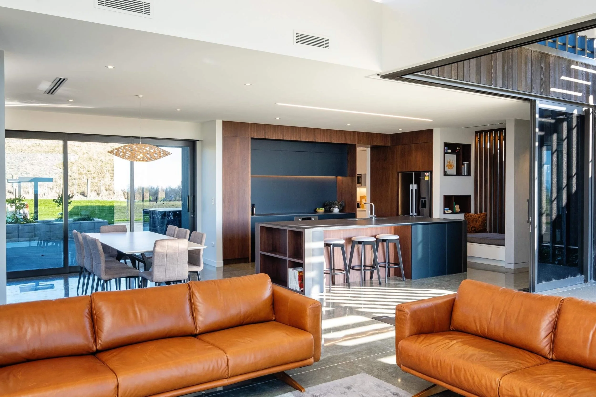 Open-concept living space with a leather sofa, a kitchen with bar stools, dining area with chairs, large sliding glass doors showing a grassy outdoor area.