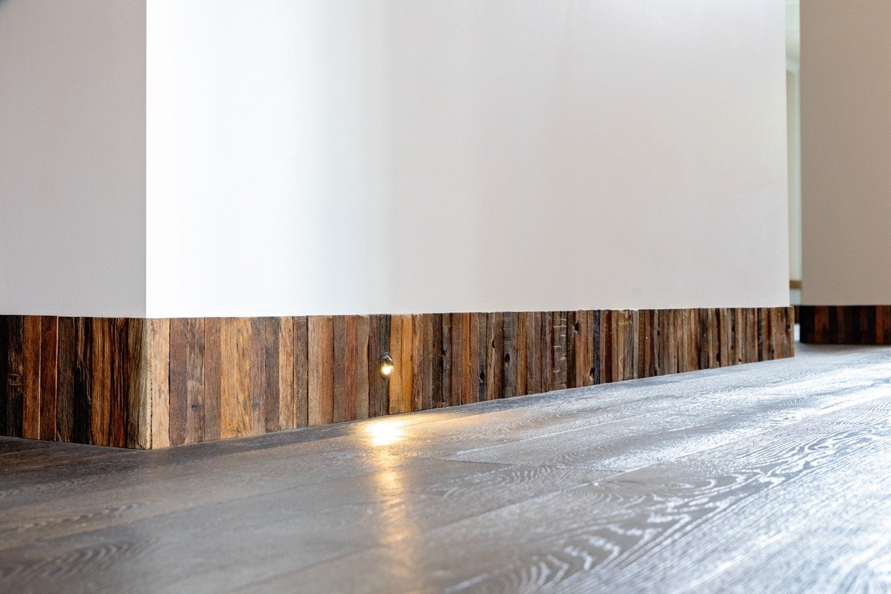 Close-up of a wooden baseboard along the bottom of a white wall, with a concealed light shining onto the floor.