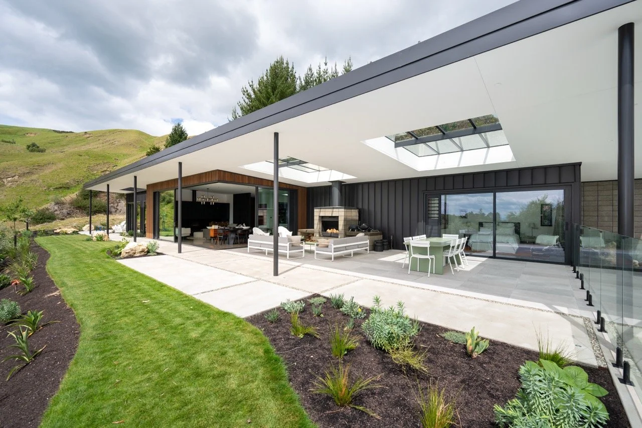 Modern house with a large outdoor patio, glass railing, outdoor fireplace, green lawn, and hillside background.