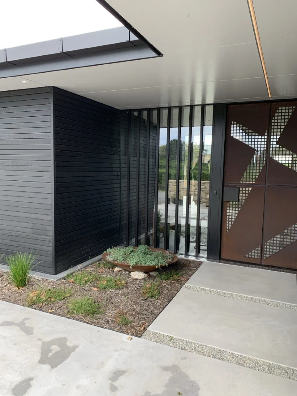 Modern building exterior with black and rust-colored metal panels, glass doors, and a small garden with plants near the entrance.