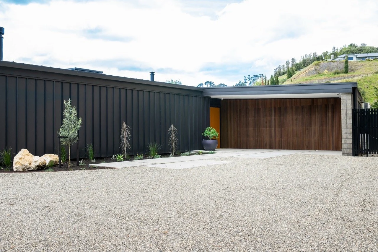 Modern house exterior with black metal and wooden siding, small plants, gravel driveway, large rock, potted plant, and hillside background.
