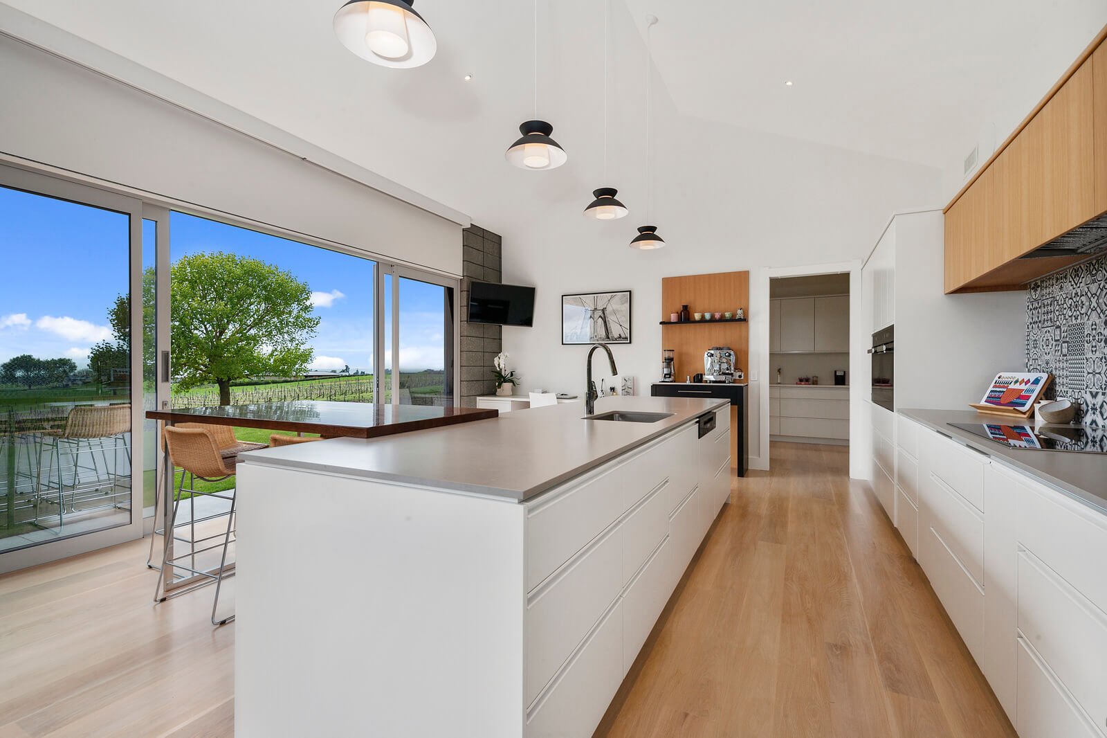 Modern kitchen with white cabinets, wooden accents, a large island with a sink, and sliding glass doors showing a tree and outdoor patio area.