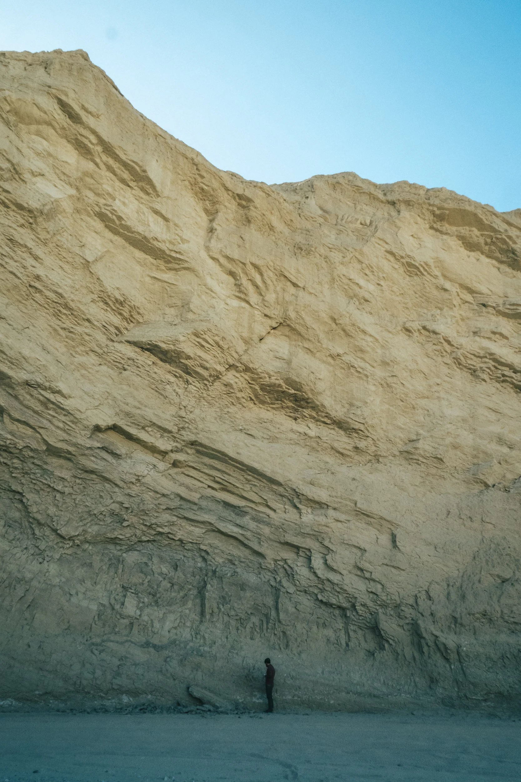 A person standing at the base of a large, beige, rocky cliff under a clear blue sky.