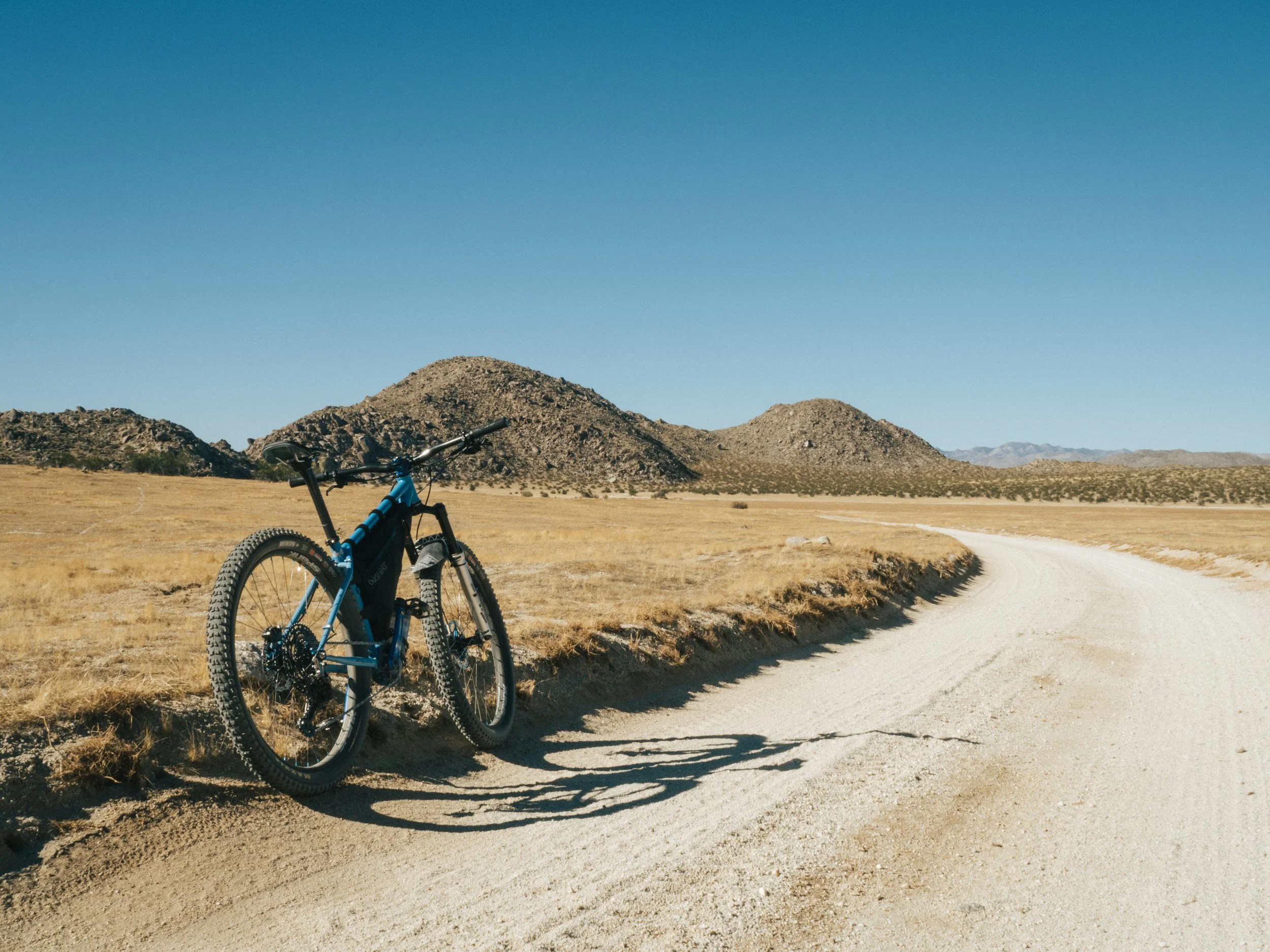 A blue mountain bike parked on a dirt road in a desert landscape with mountains in the background and a clear blue sky.