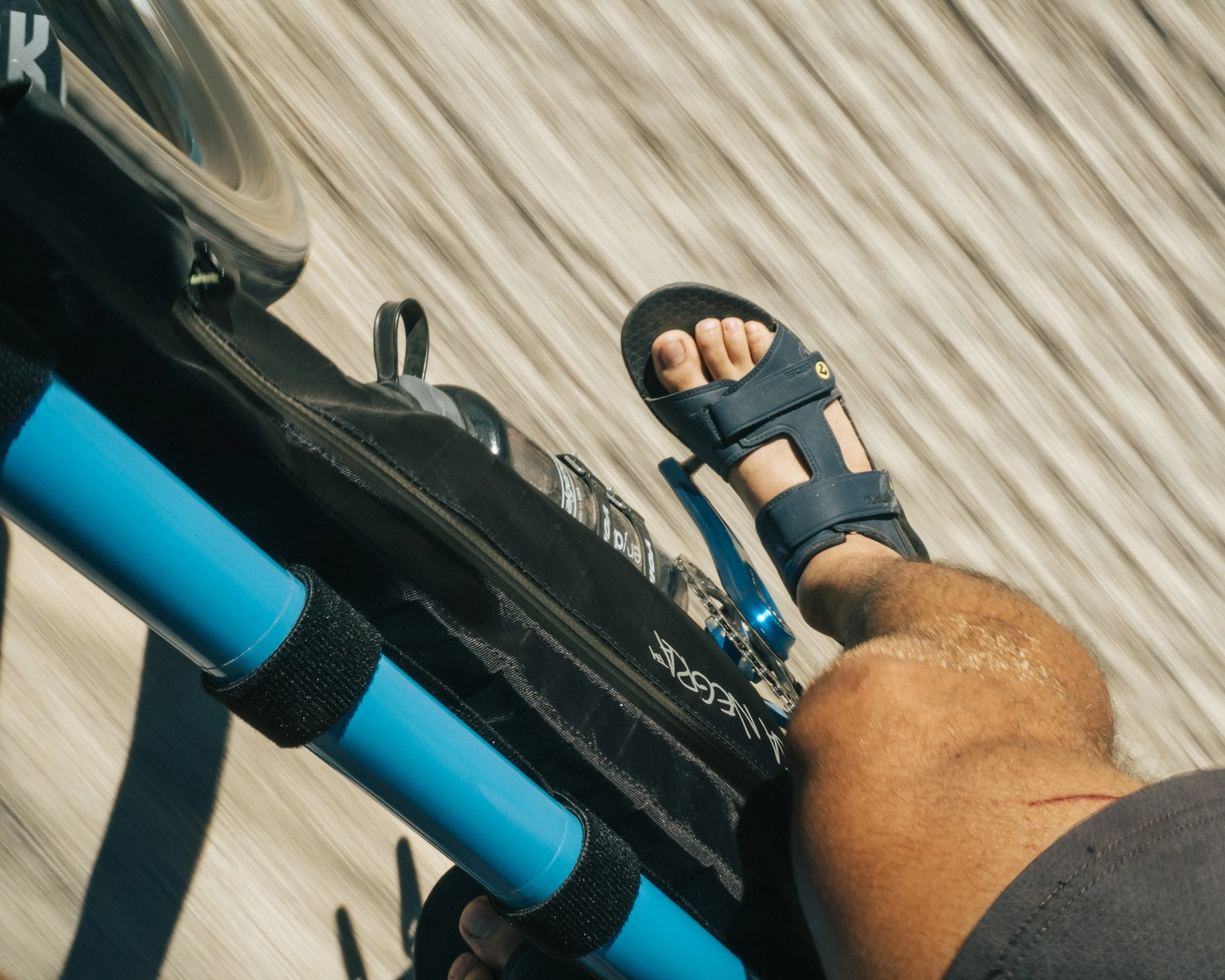Close-up of a person's leg with a sandal on a skateboard with a black carrying case and water bottle, moving on a wooden surface.