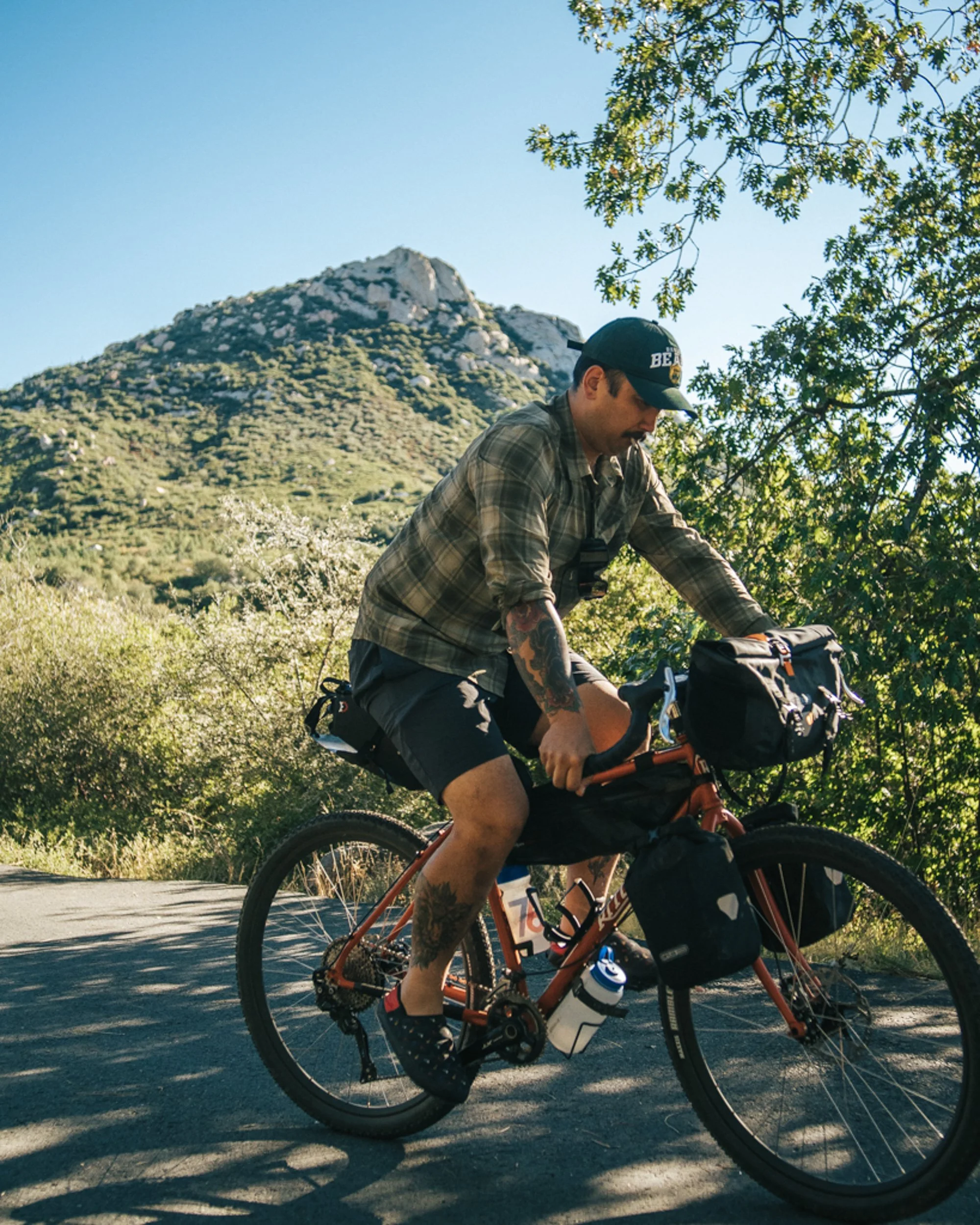 Man riding a bicycle on a paved trail with mountains and trees in the background.