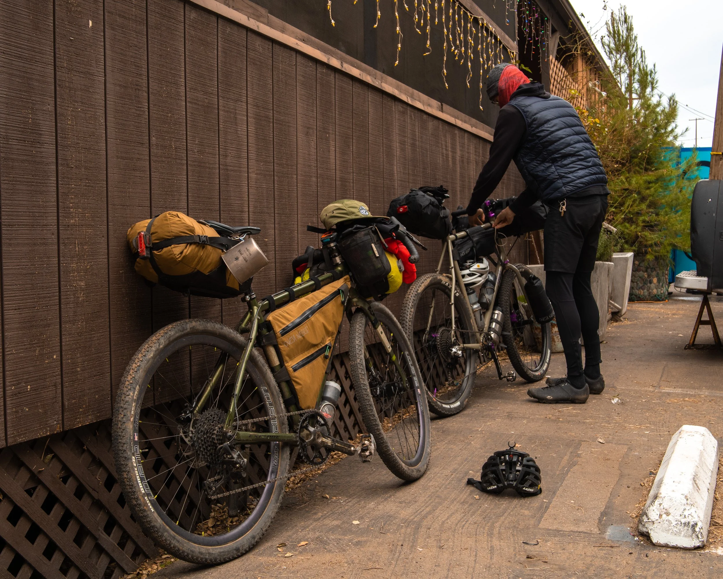 A person in outdoor gear adjusting their bike with multiple bags, standing next to two loaded bicycles leaning against a wooden wall, with a bicycle helmet on the ground nearby.
