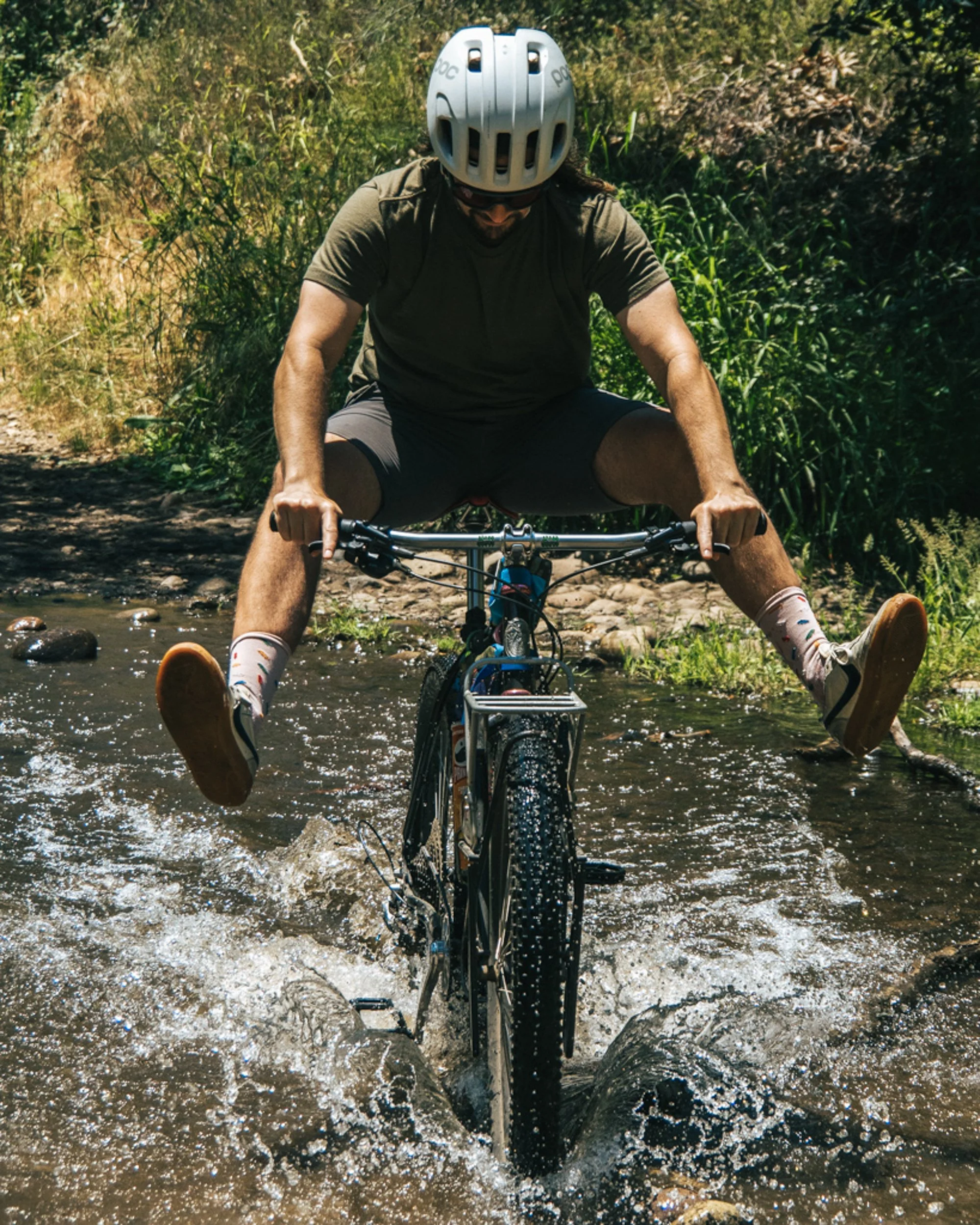 A man riding a bicycle through a shallow stream in a forested area, wearing a helmet, sunglasses, a green T-shirt, and shorts.