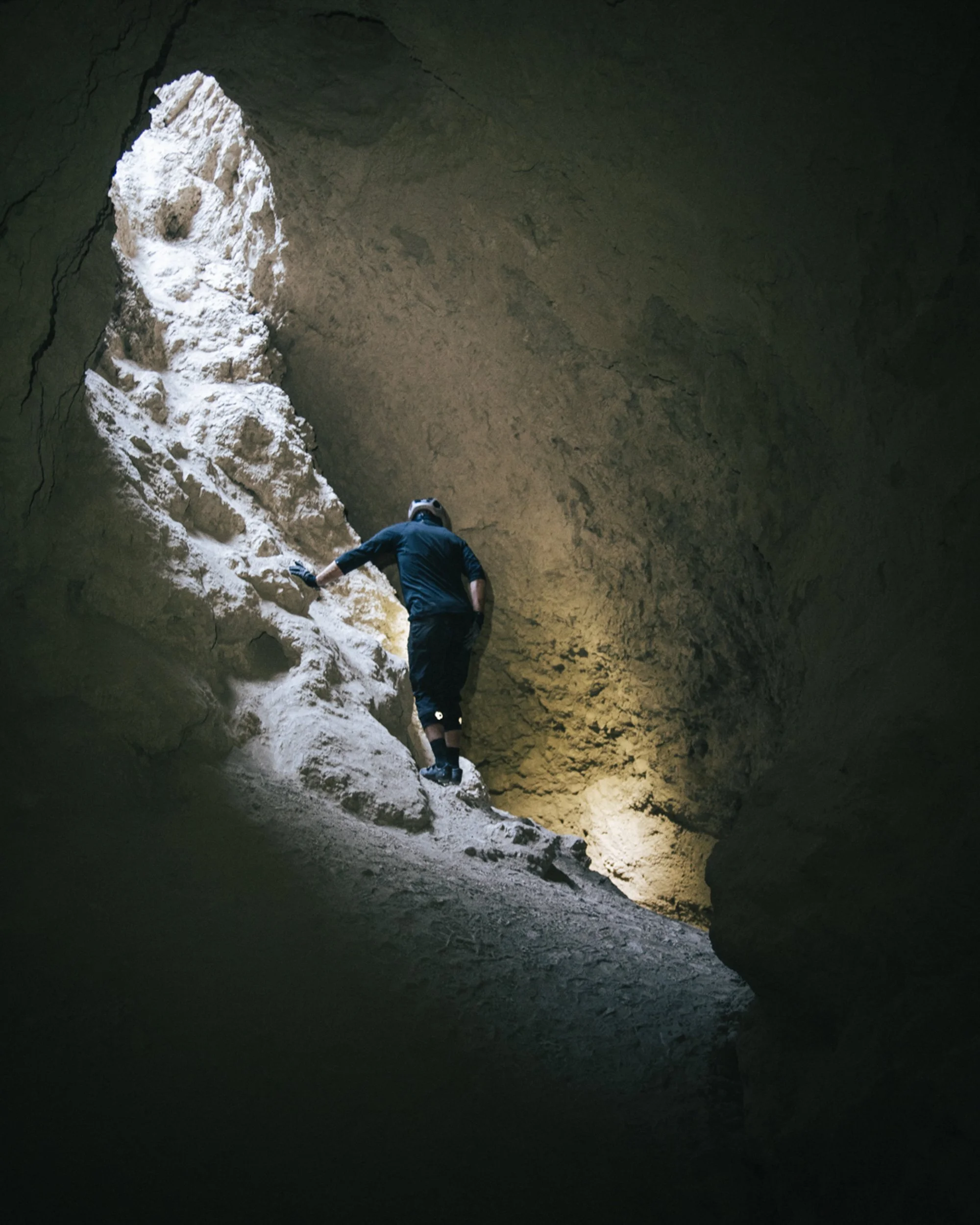Person wearing a helmet and dark clothing exploring inside a cave, climbing over rocks with natural light illuminating the rocky walls.
