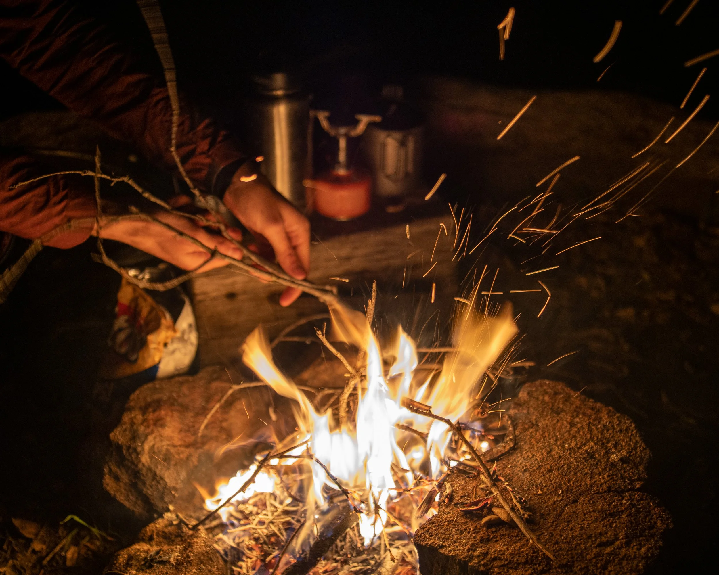 Person tending a campfire surrounded by rocks, with camping gear in the background at night.