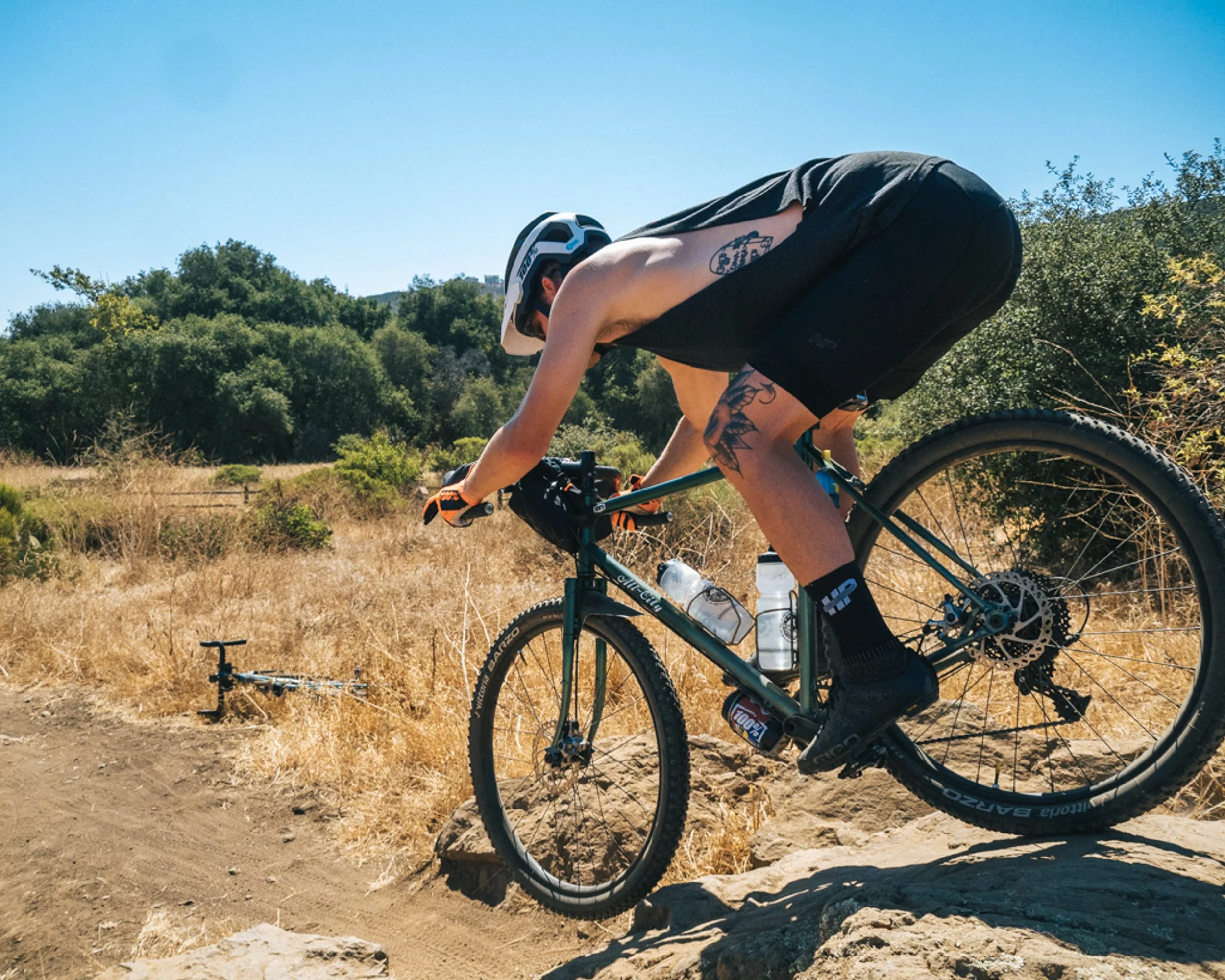 A person in a black sleeveless shirt and shorts riding a mountain bike downhill on a rocky trail, with a helmet and water bottles attached to the bike, in a dry, grassy landscape with trees under a clear blue sky.
