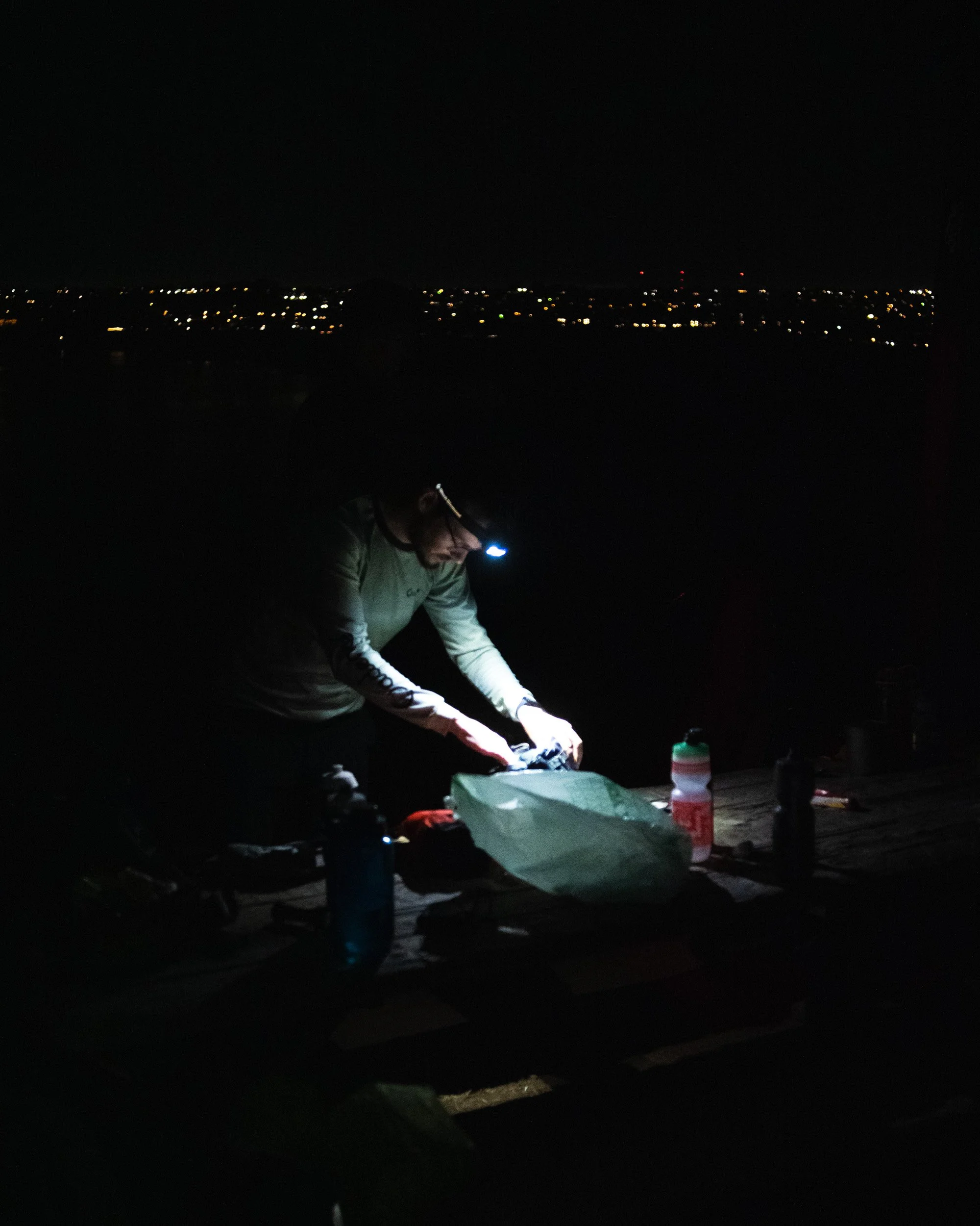 Person with headlamp arranging camping gear at night, with a dark lake and city lights in the distance.