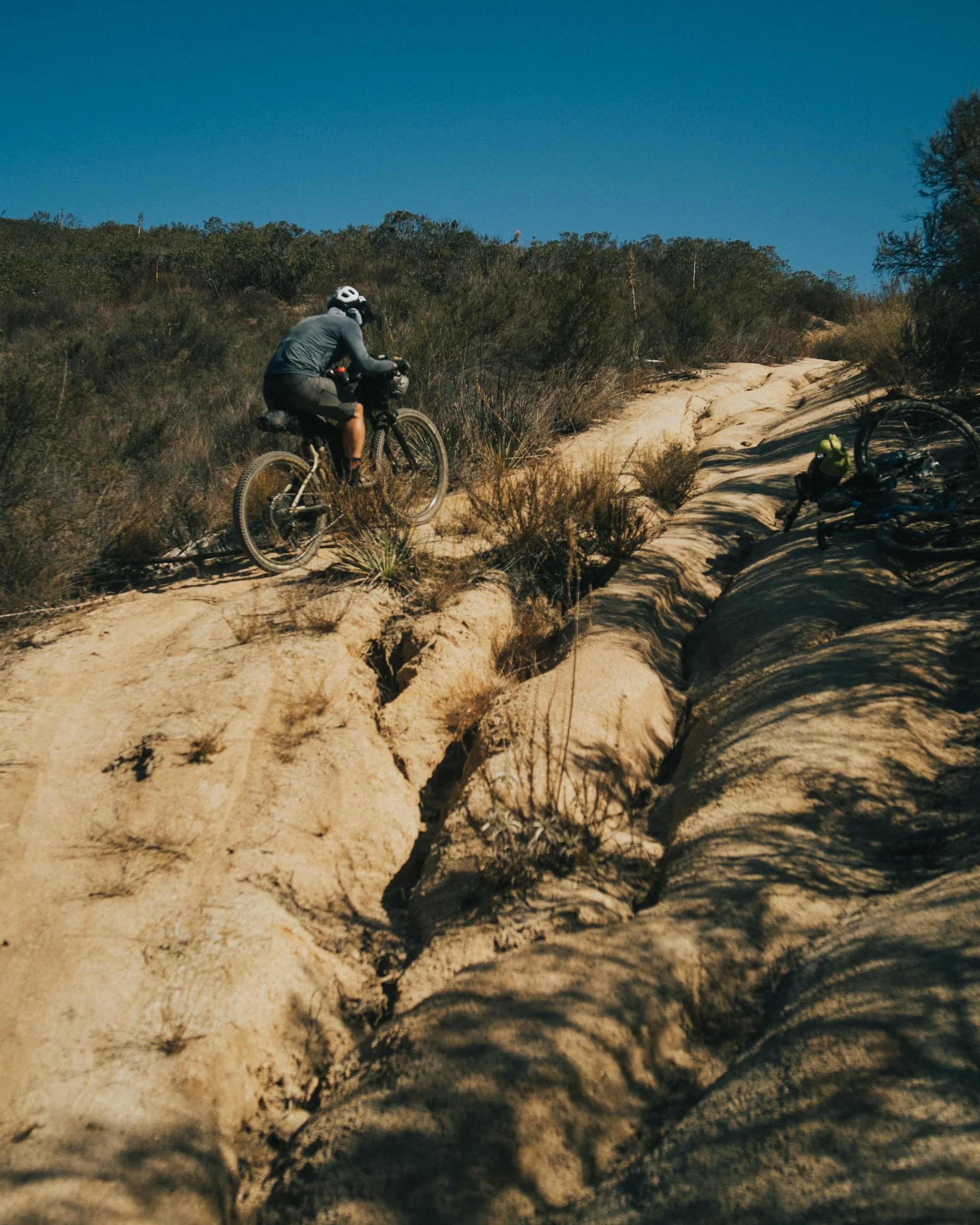 A person riding a mountain bike up a dirt trail on a sunny day, with another bike lying on the ground to the side and dry bushes around.