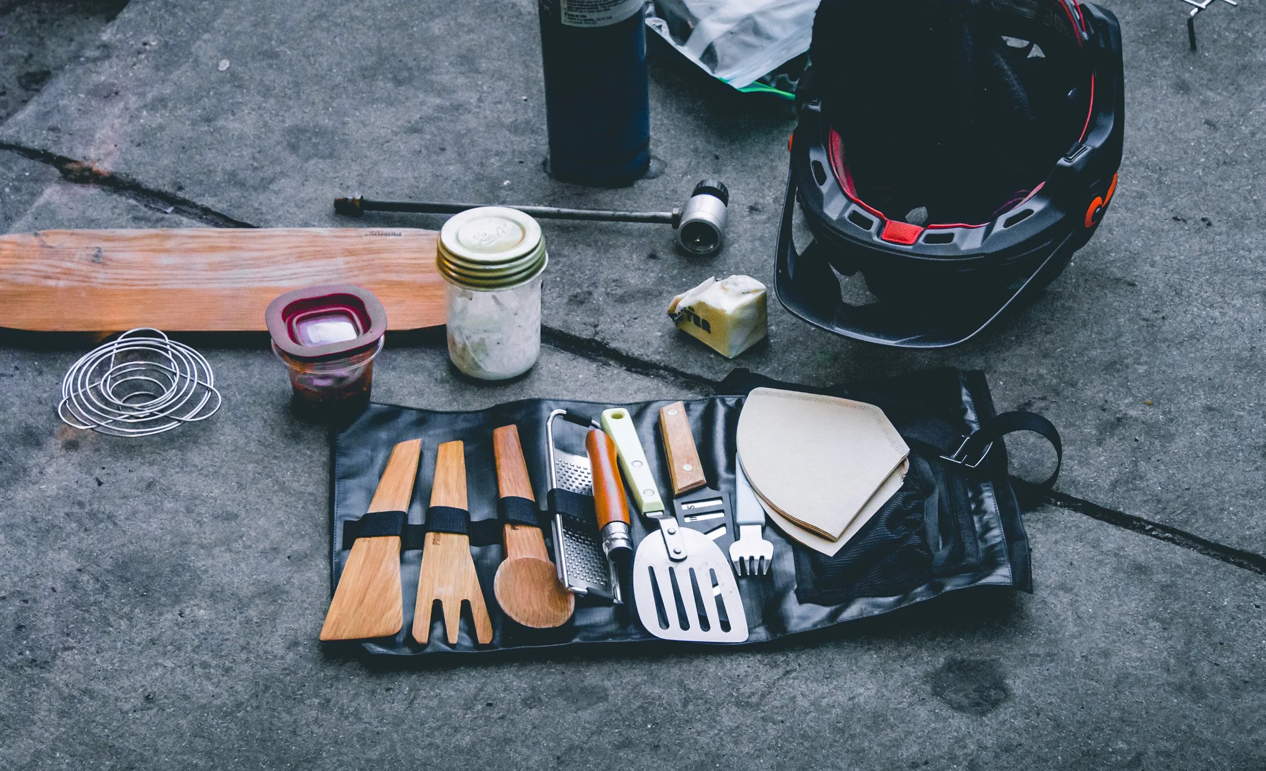 Set of barbecue tools with wooden handles, including spatula, fork, and tongs, laid out on a black mat on a concrete surface. Nearby are a helmet, mason jar, small containers, a wooden plank, and a flashlight.