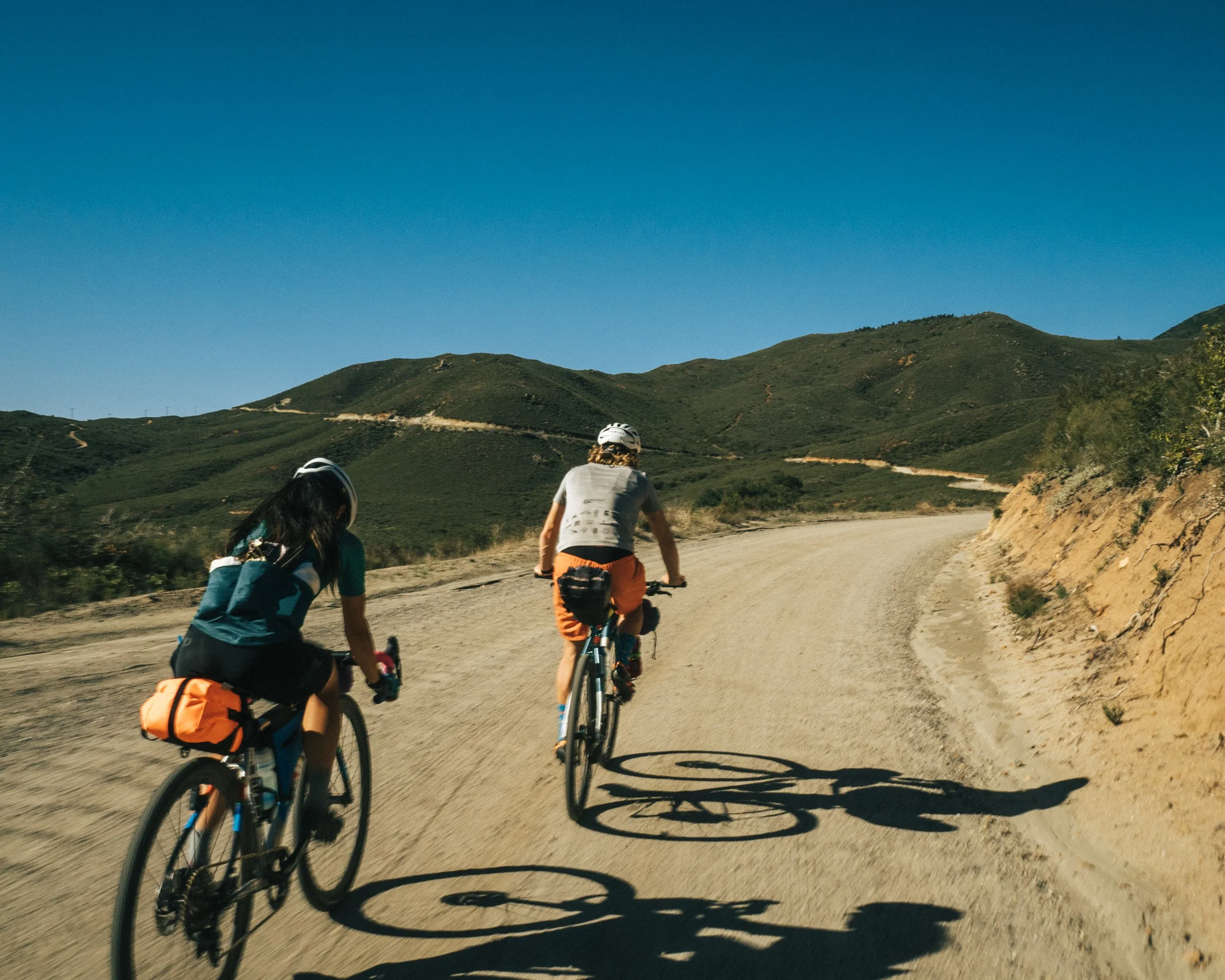 Two people mountain biking on a dirt trail in a hilly landscape under a clear blue sky.