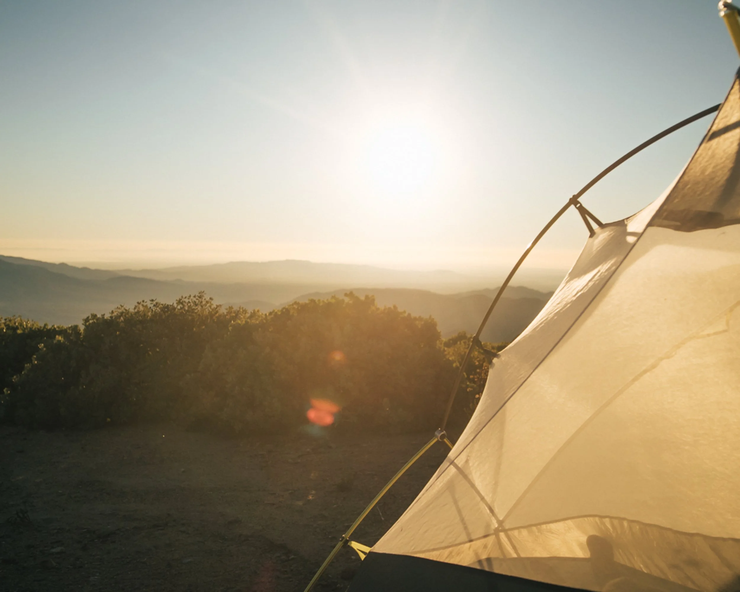 A camping tent set up on a hilltop with a scenic view of rolling mountains and bushes at sunset or sunrise.