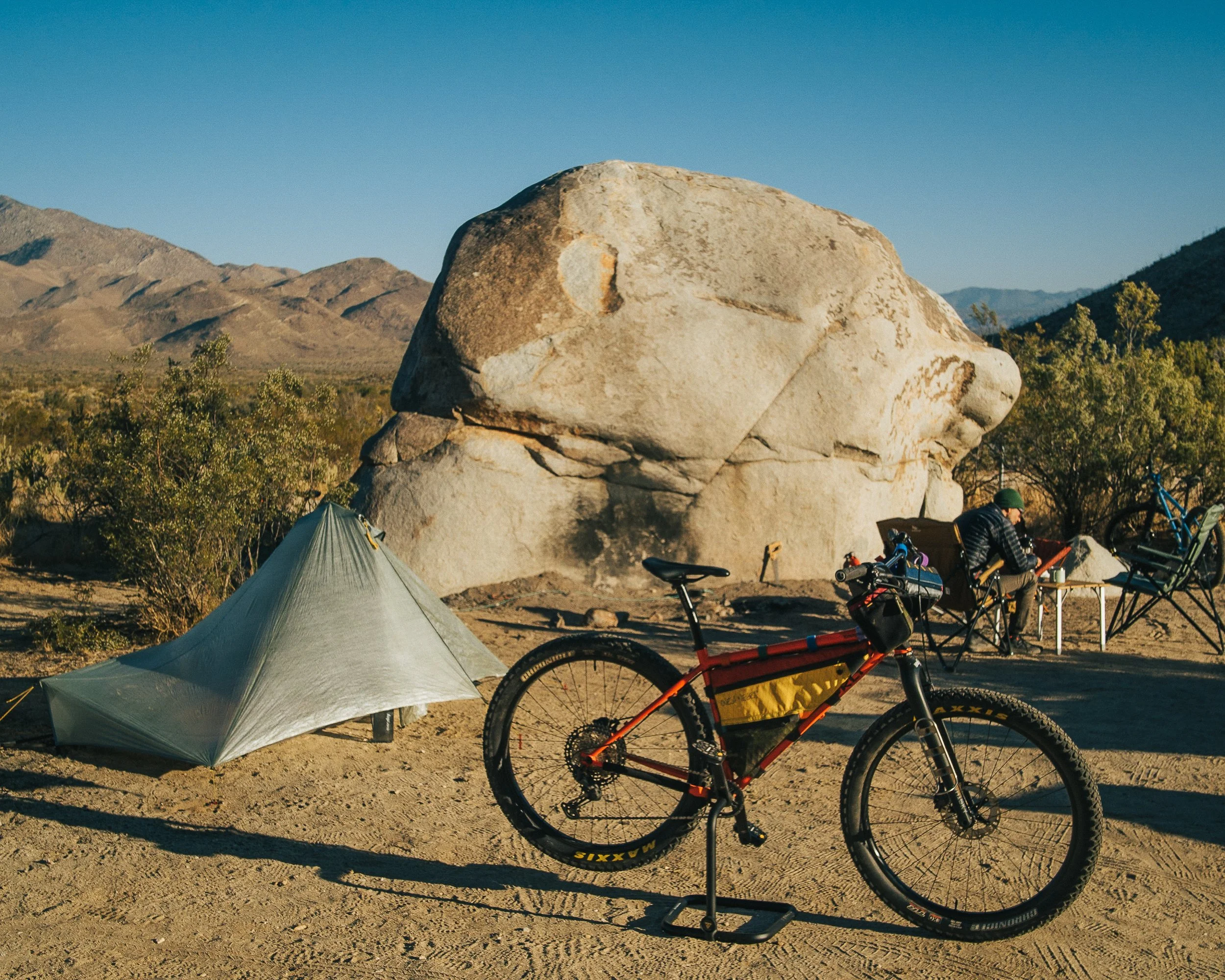 A desert campsite with a tent, mountain bike, and a person sitting at a table near a large boulder, with mountains in the background under a clear sky.