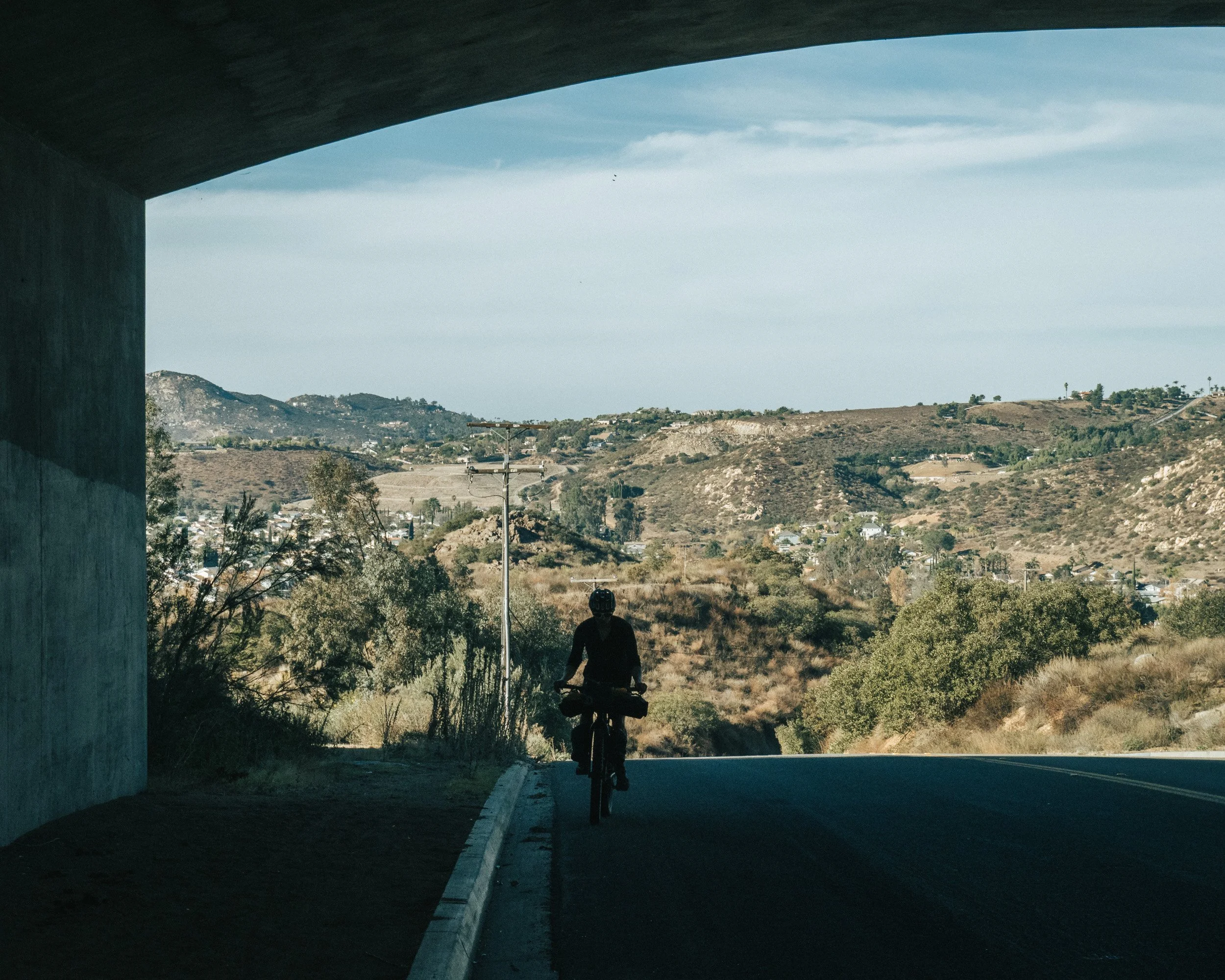 A person riding a bicycle under a bridge with hilly terrain and scattered trees in the background.