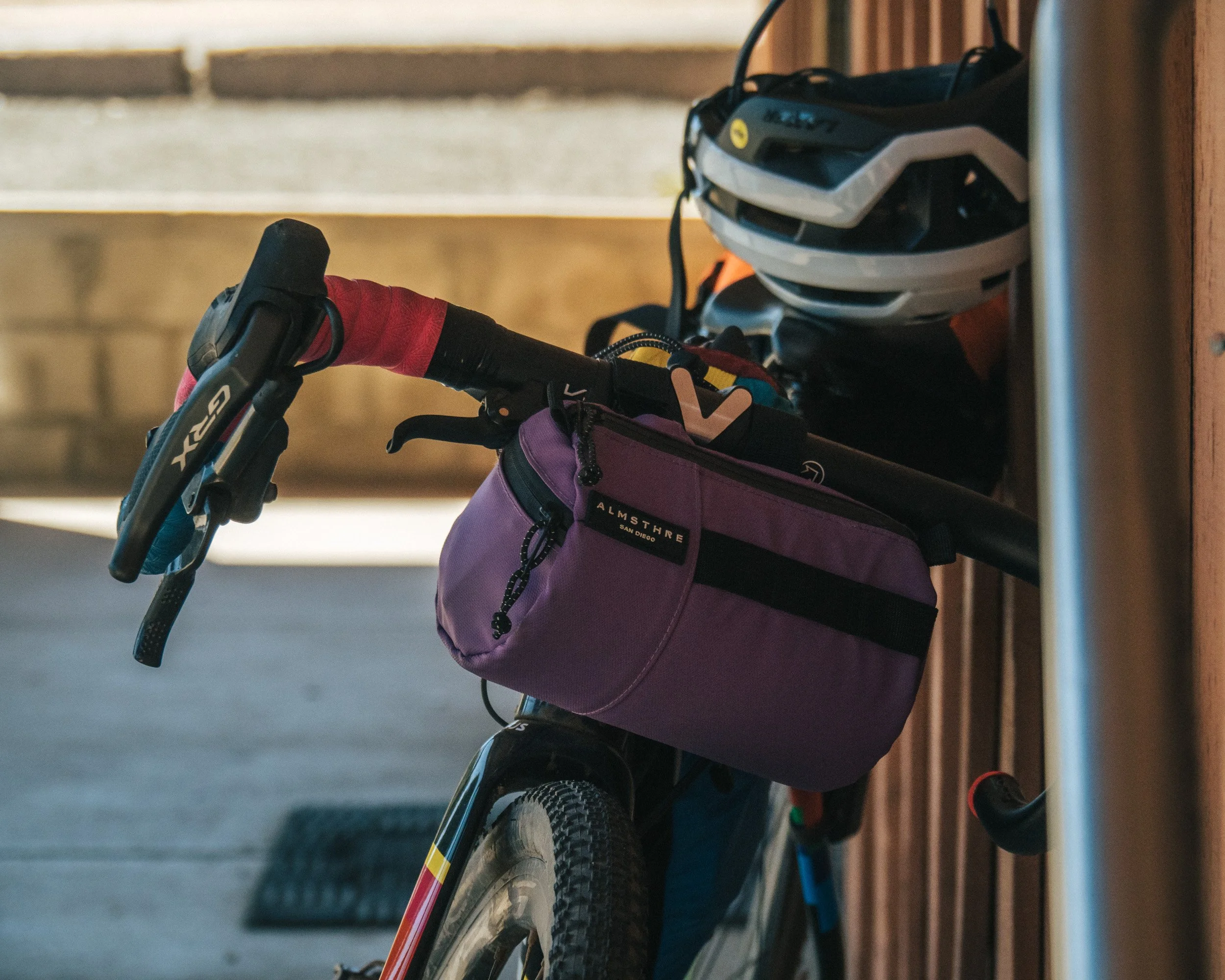 A mountain bike with a purple bag hanging from the handlebar, a white helmet on the frame, and a black headlamp, leaning against a wooden wall.