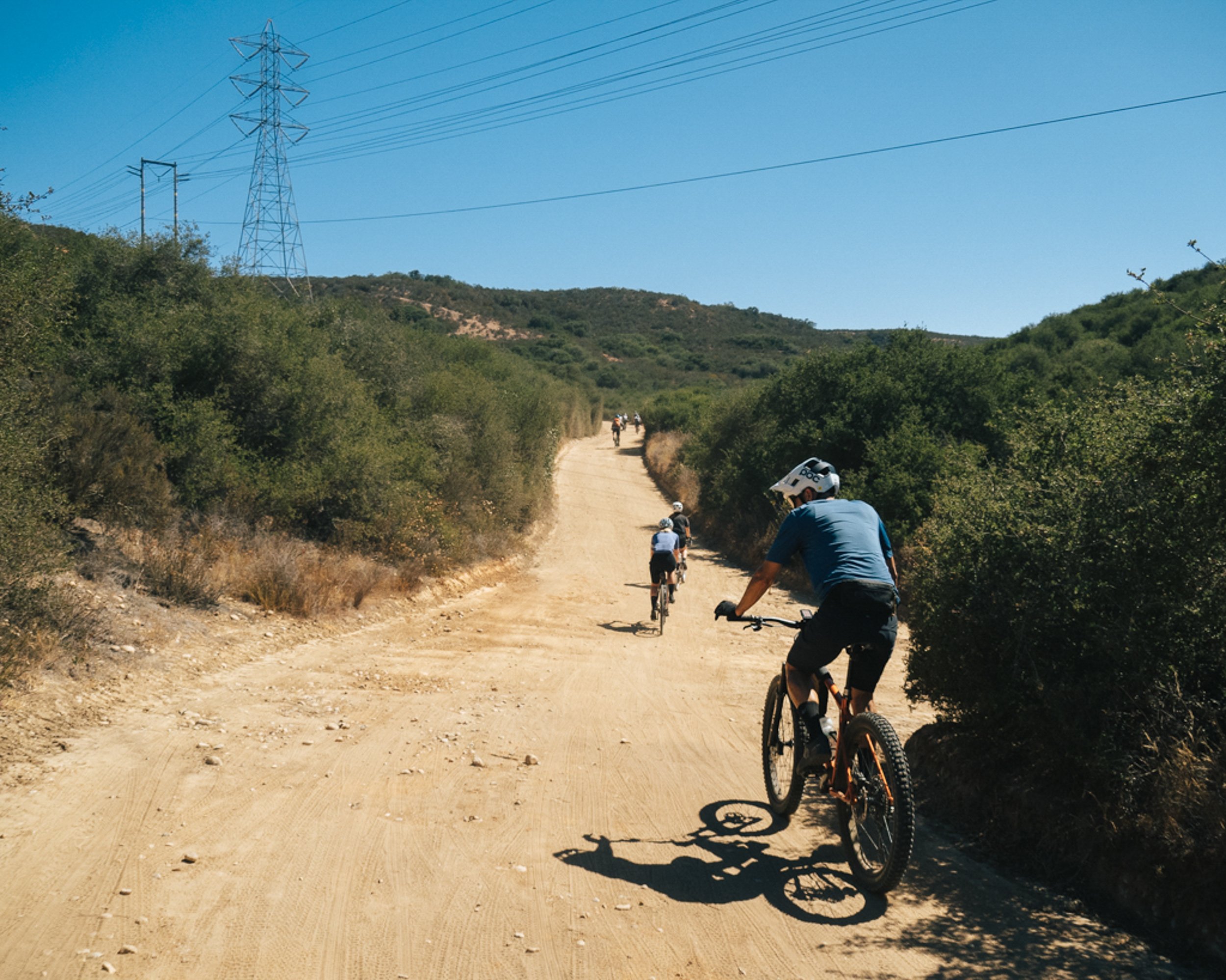 Group of cyclists riding mountain bikes on an unpaved trail through a hilly, green landscape with power lines and a clear blue sky.