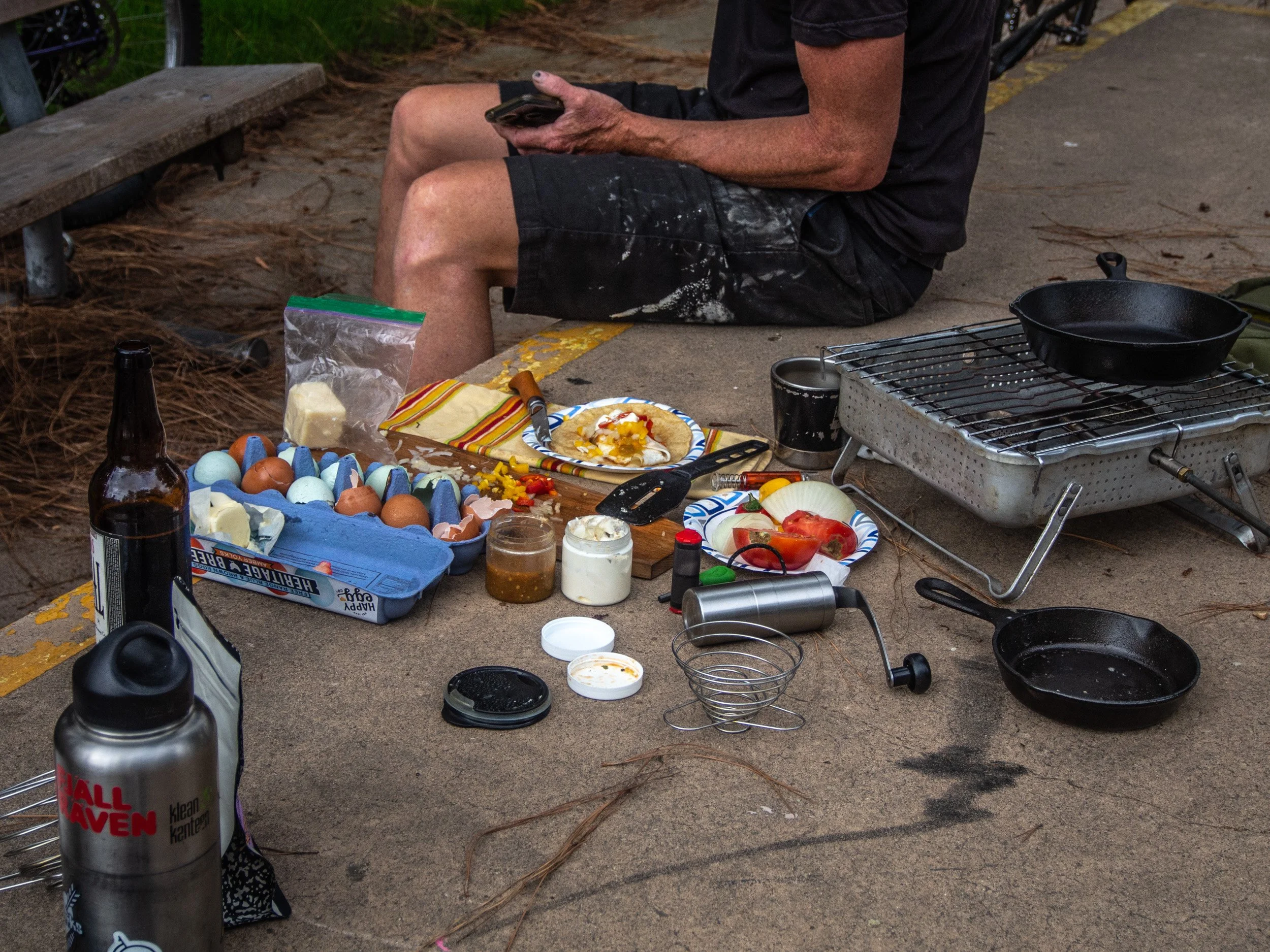 Outdoor picnic table with food and cooking supplies, including eggs, a bottle, plates, jars, a pan, and a man sitting nearby.