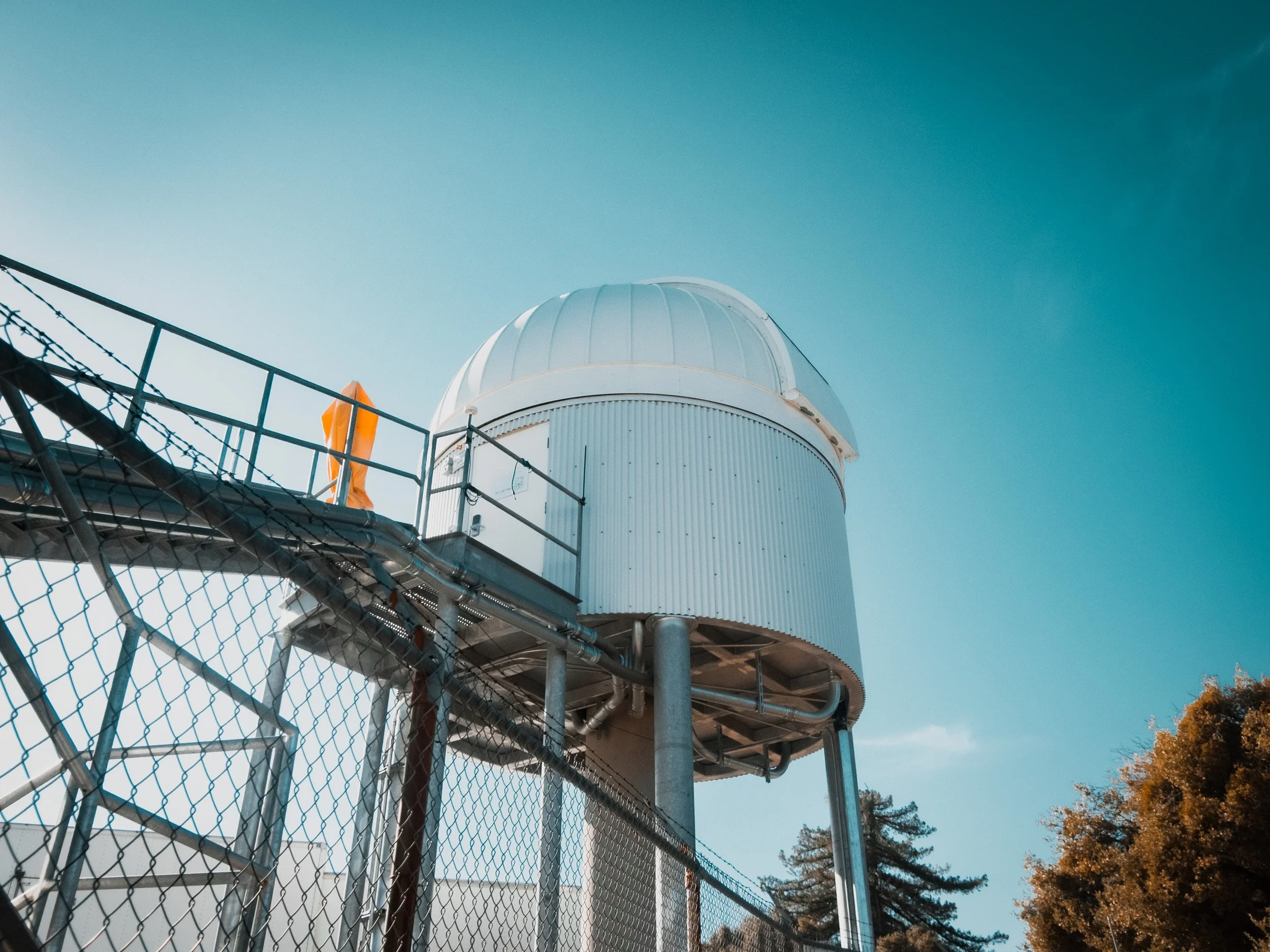 An observatory dome on a tall structure with a chain-link fence in front, under a blue sky with some clouds and trees at the bottom right corner.