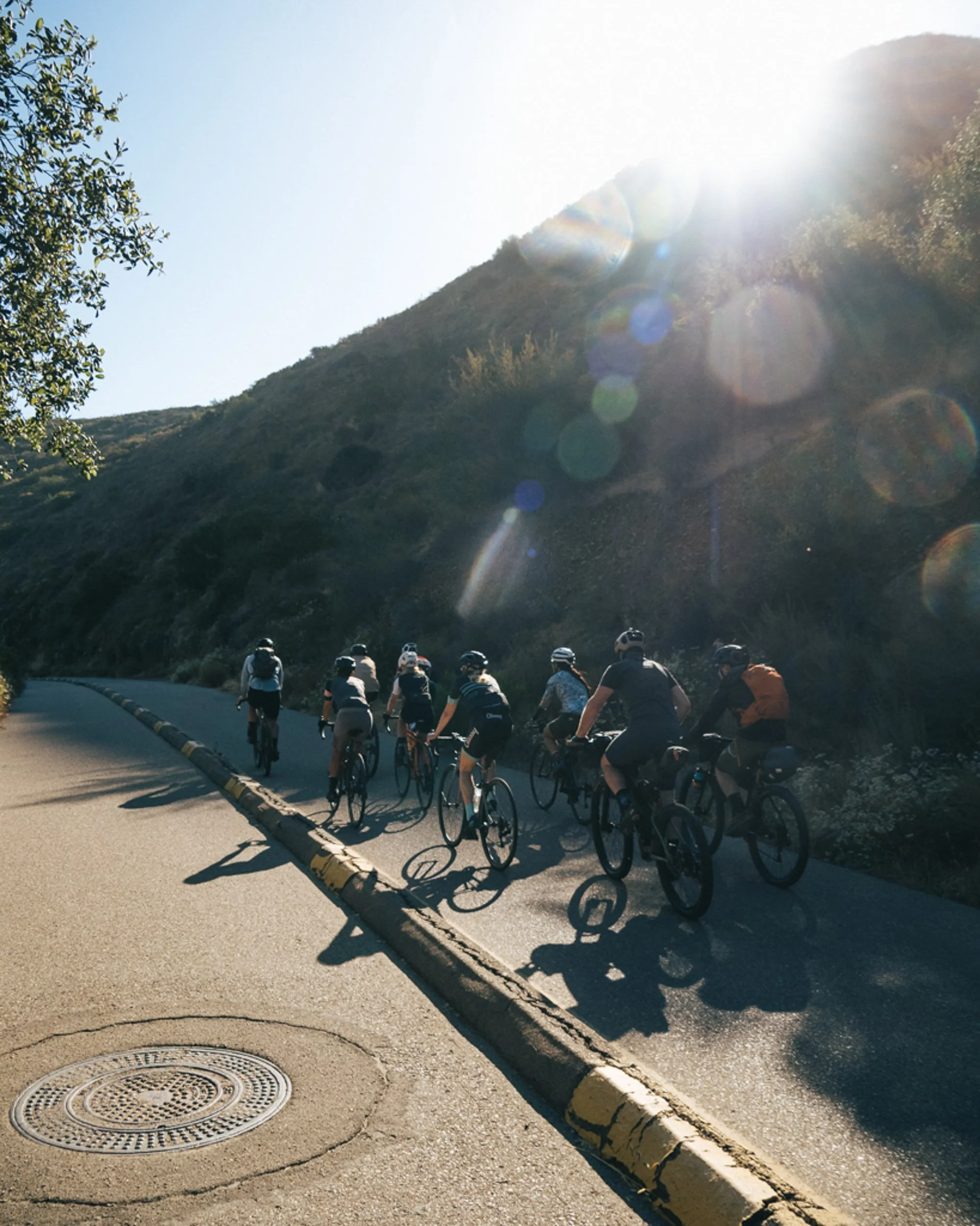 A group of cyclists riding uphill on a paved road in a mountainous area with the sun shining brightly overhead, creating lens flare.