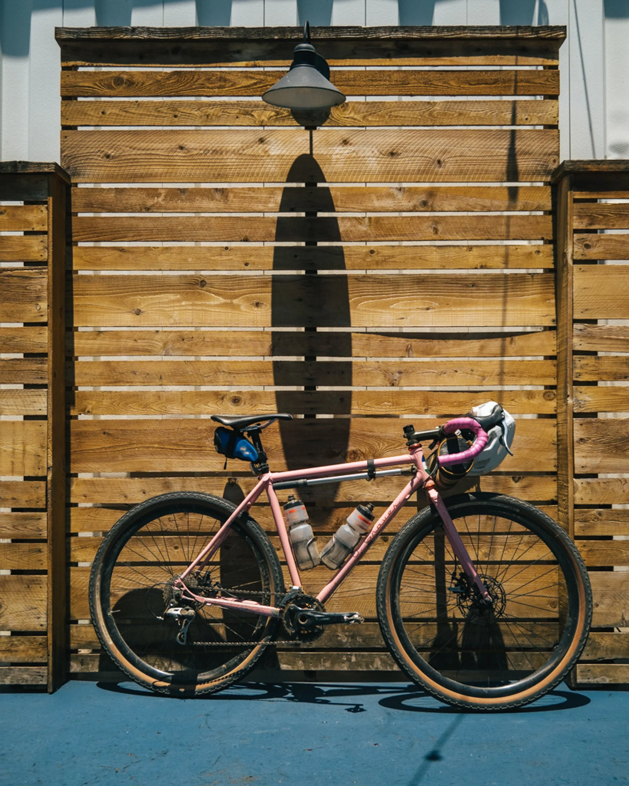 A pink mountain bike with water bottles attached, leaning against a wooden fence outdoor, with a shadow of a street lamp cast on the fence.