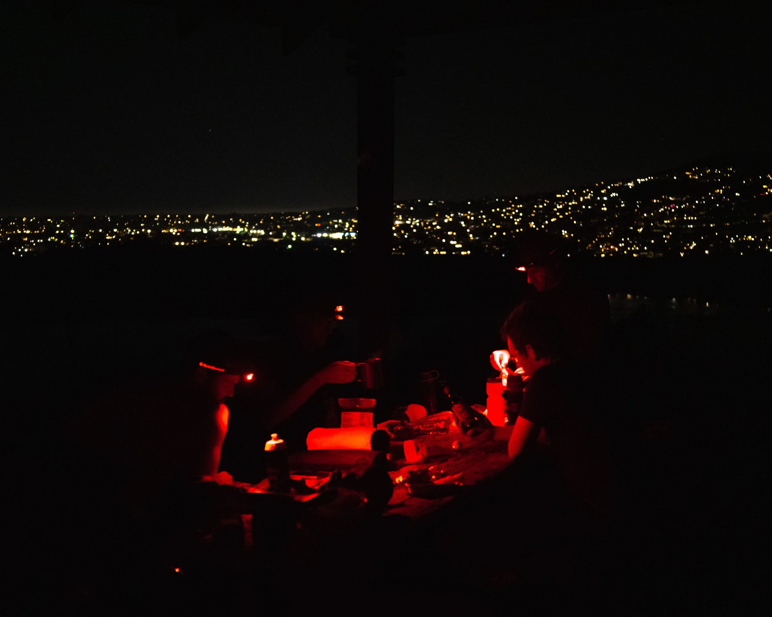 People sitting at a table outdoors at night, illuminated by red lights, with a cityscape of lights in the background.