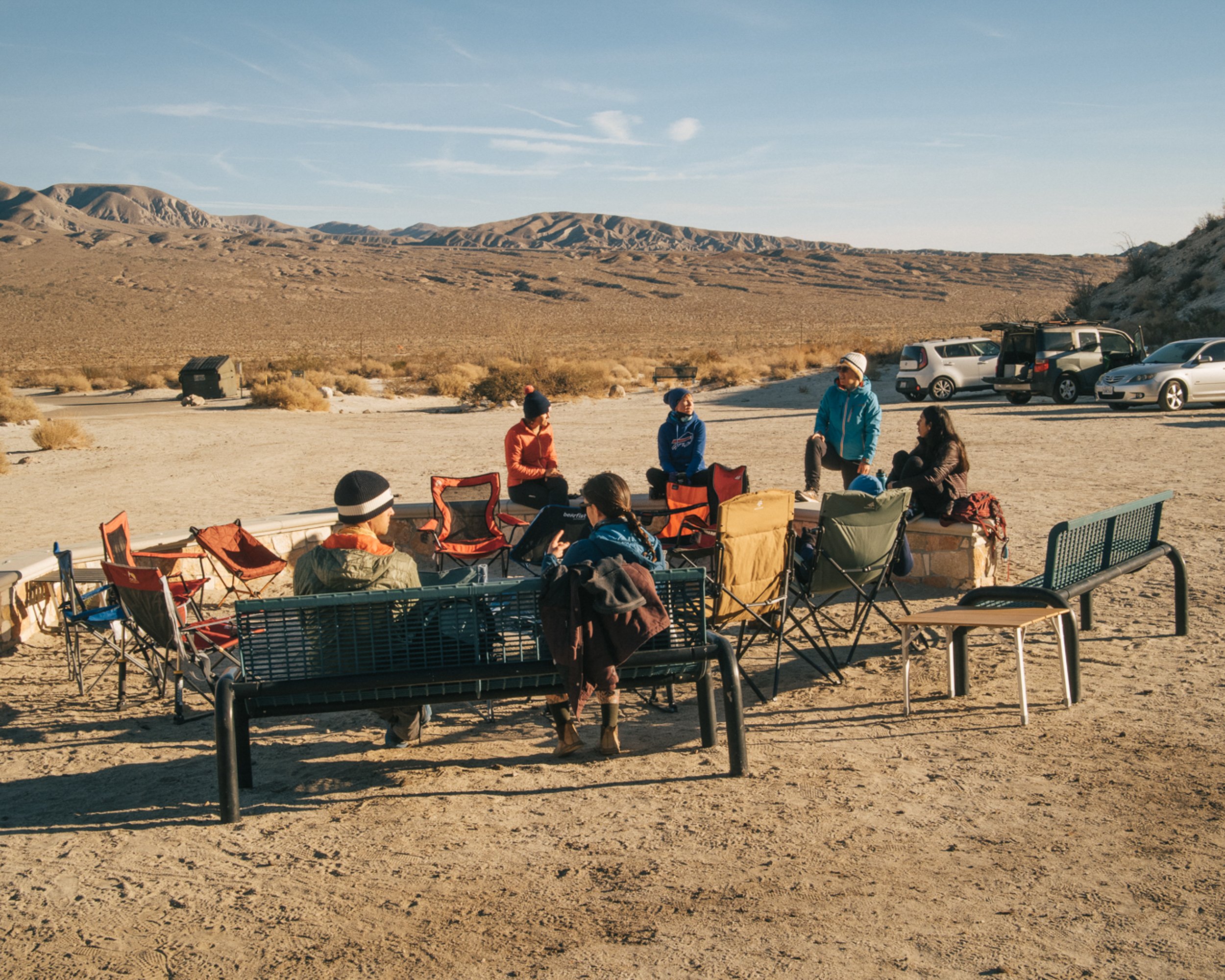 A group of people gathered outdoors in a desert landscape with mountains in the background, sitting on chairs and benches, some standing, near parked cars.