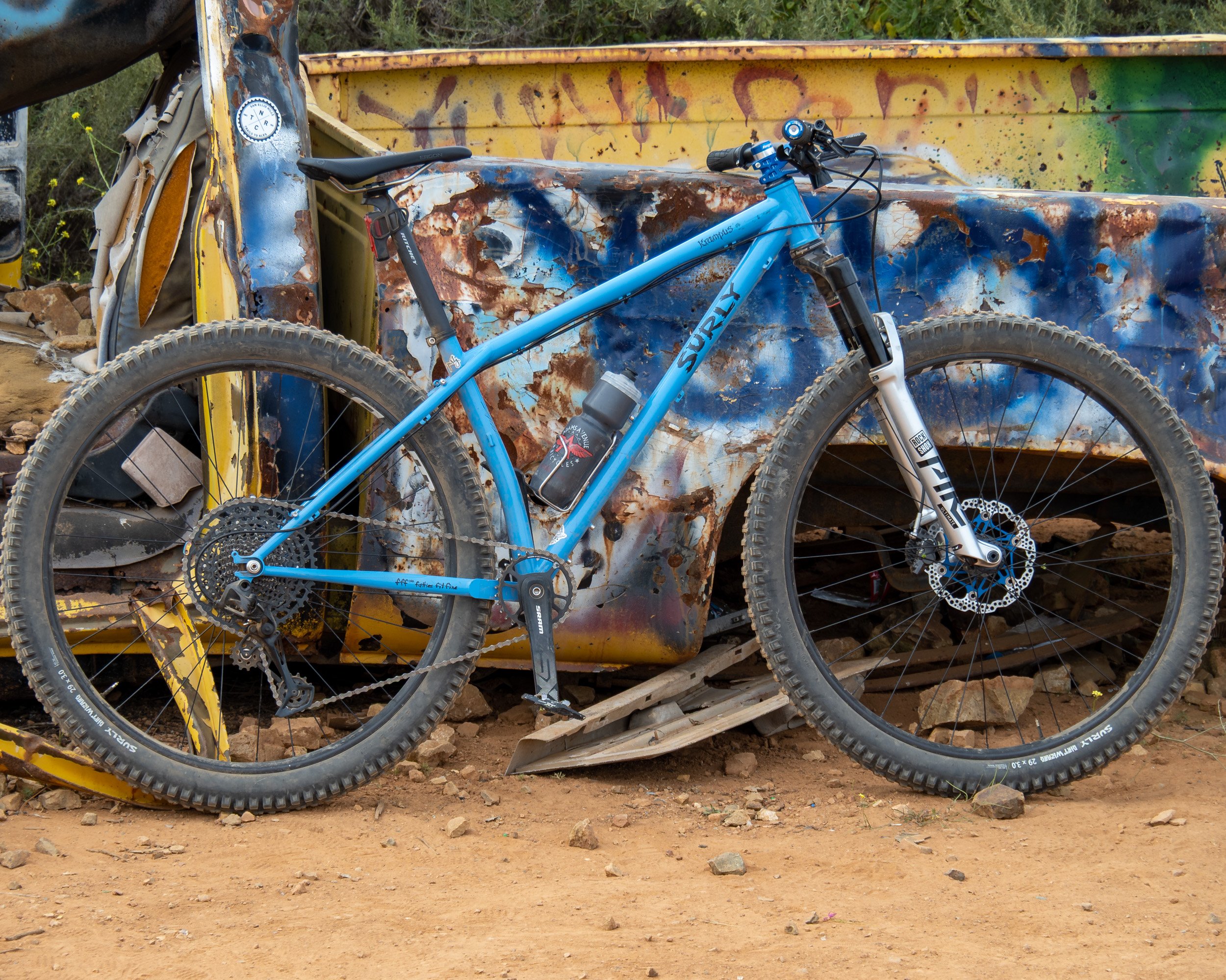 Blue mountain bike leaning against a rusted, graffiti-covered metal structure on a dirt surface.
