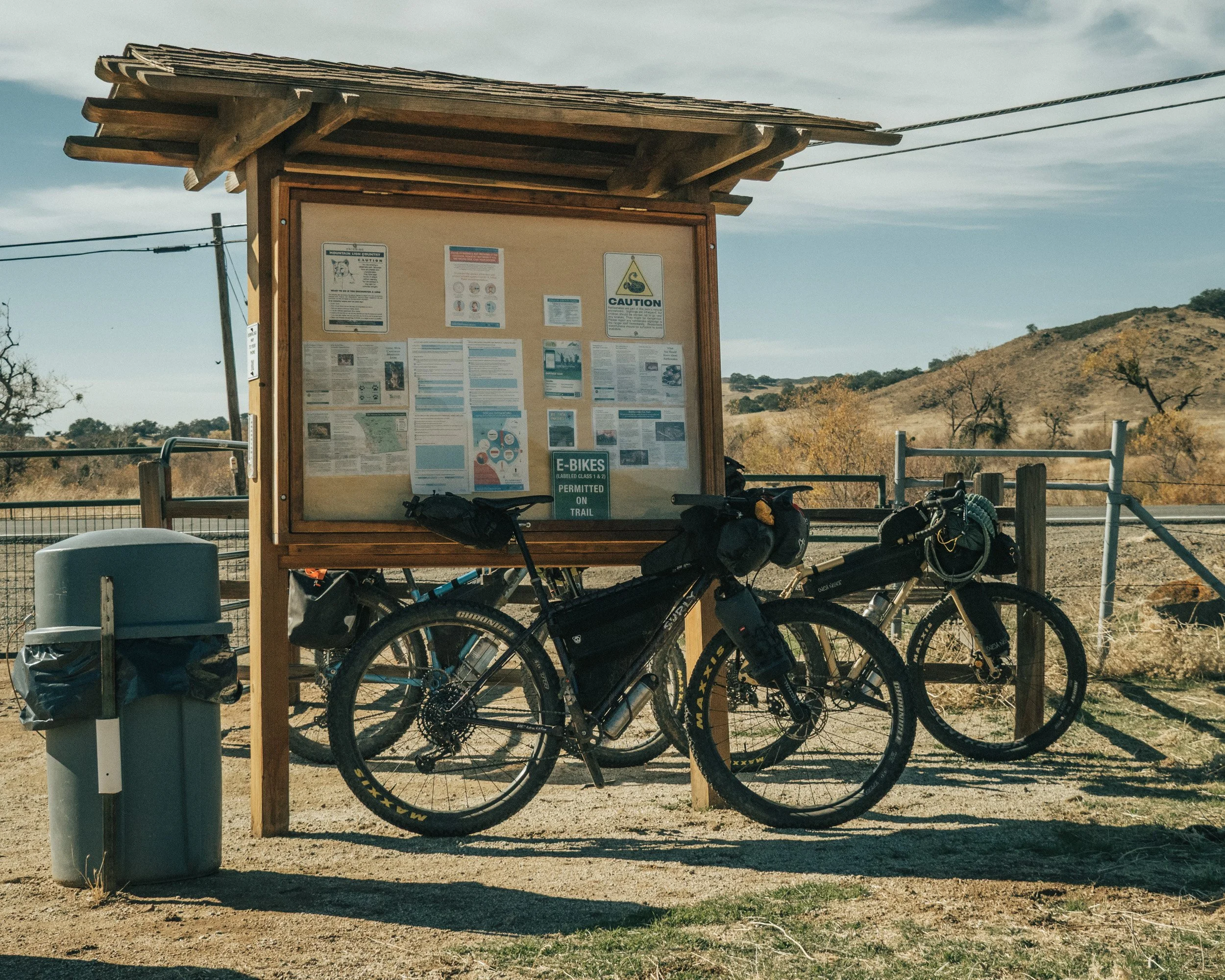 Two bicycles parked at a trailhead with an informational sign and a trash can, in a dry outdoor landscape with hills and sparse trees in the background.