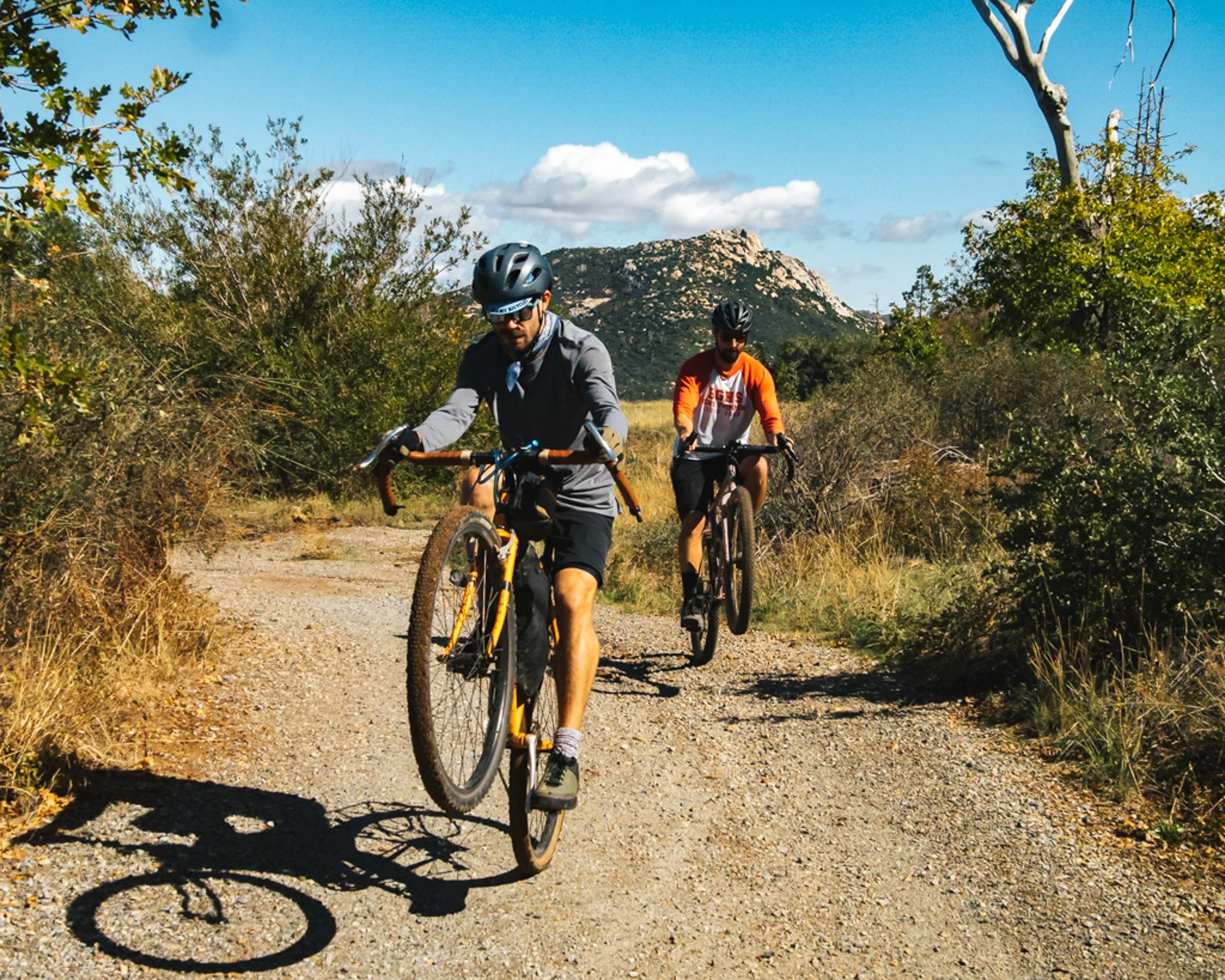 Two men riding mountain bikes on a dirt trail through a dry, green, and hilly landscape with mountains in the background under a partly cloudy sky.