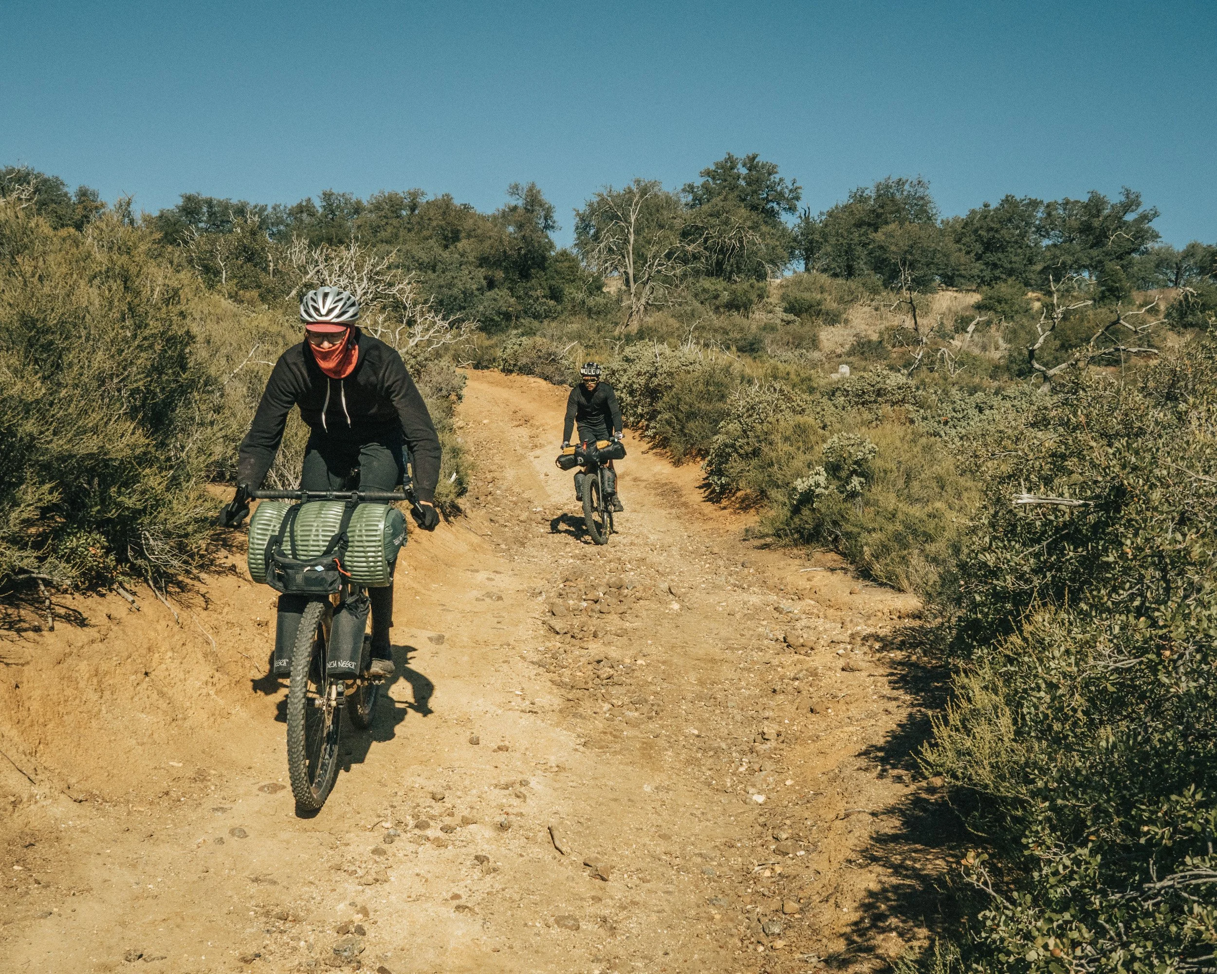 Two cyclists riding mountain bikes on a dirt trail through a dry, shrub-covered landscape under a clear blue sky.