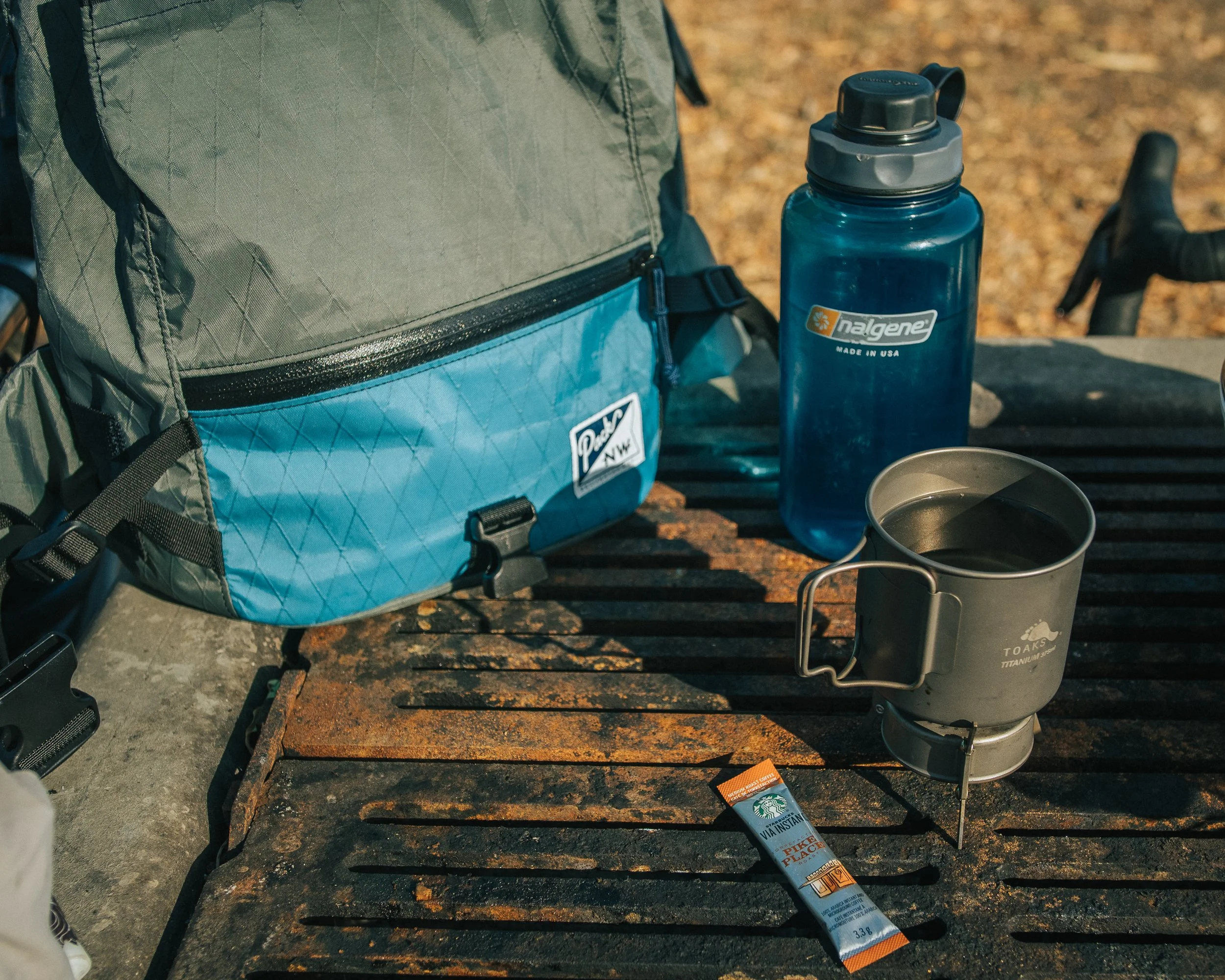 Outdoor camping scene with a blue backpack, Nalgene water bottle, titanium mug, and a small packet of Starbucks VIA instant coffee on a rusty metal surface with rocks and leaves in the background.