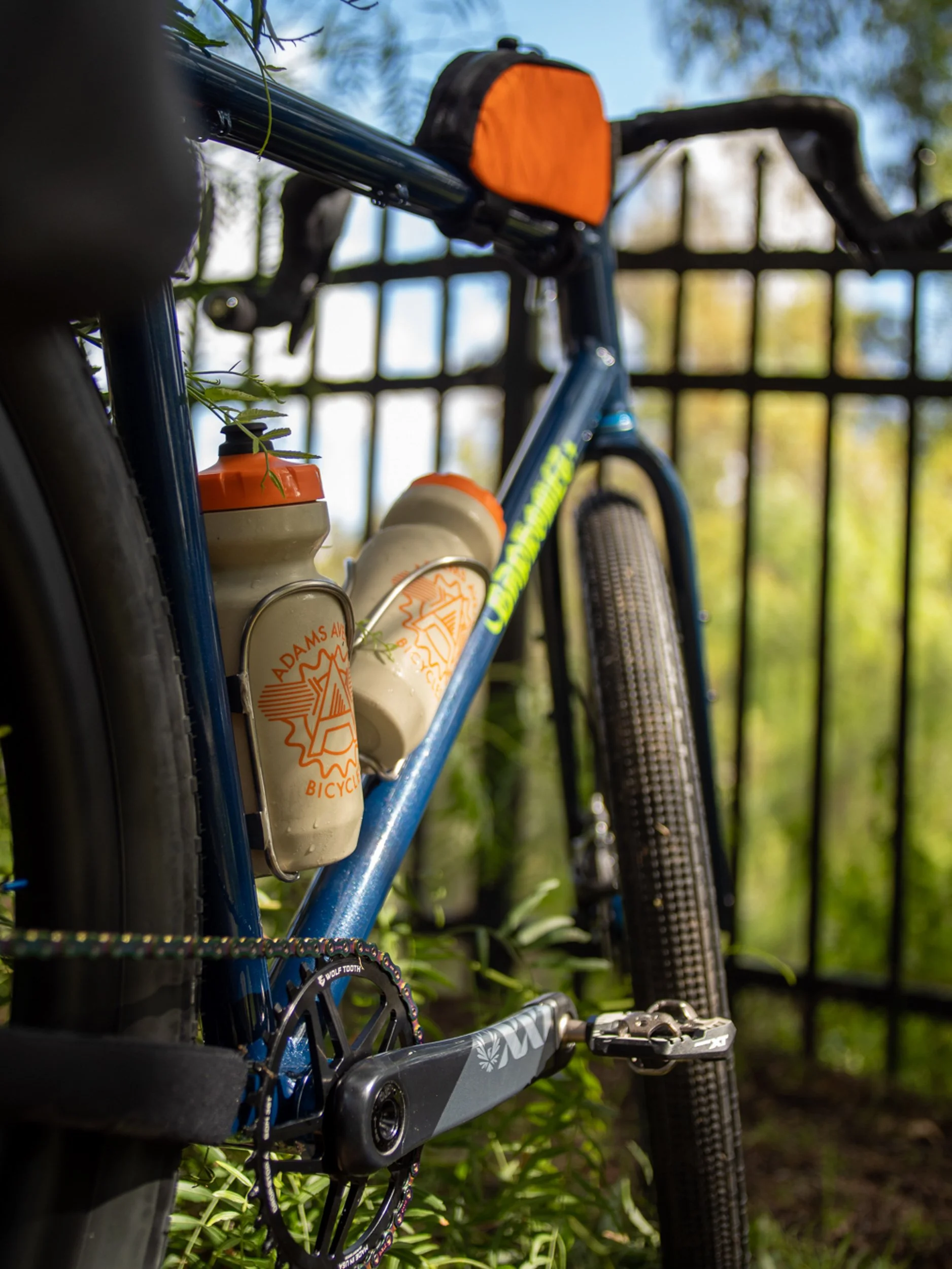 Close-up of a blue mountain bike with two beige water bottles attached to the frame, set against a backdrop of green foliage and a black metal fence.