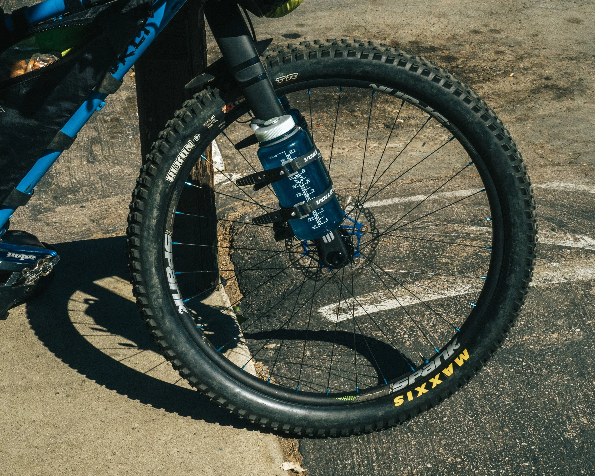Close-up of a mountain bike wheel with Maxxis tires, a suspension fork, and water bottles attached to the frame, parked on asphalt with white parking lines.